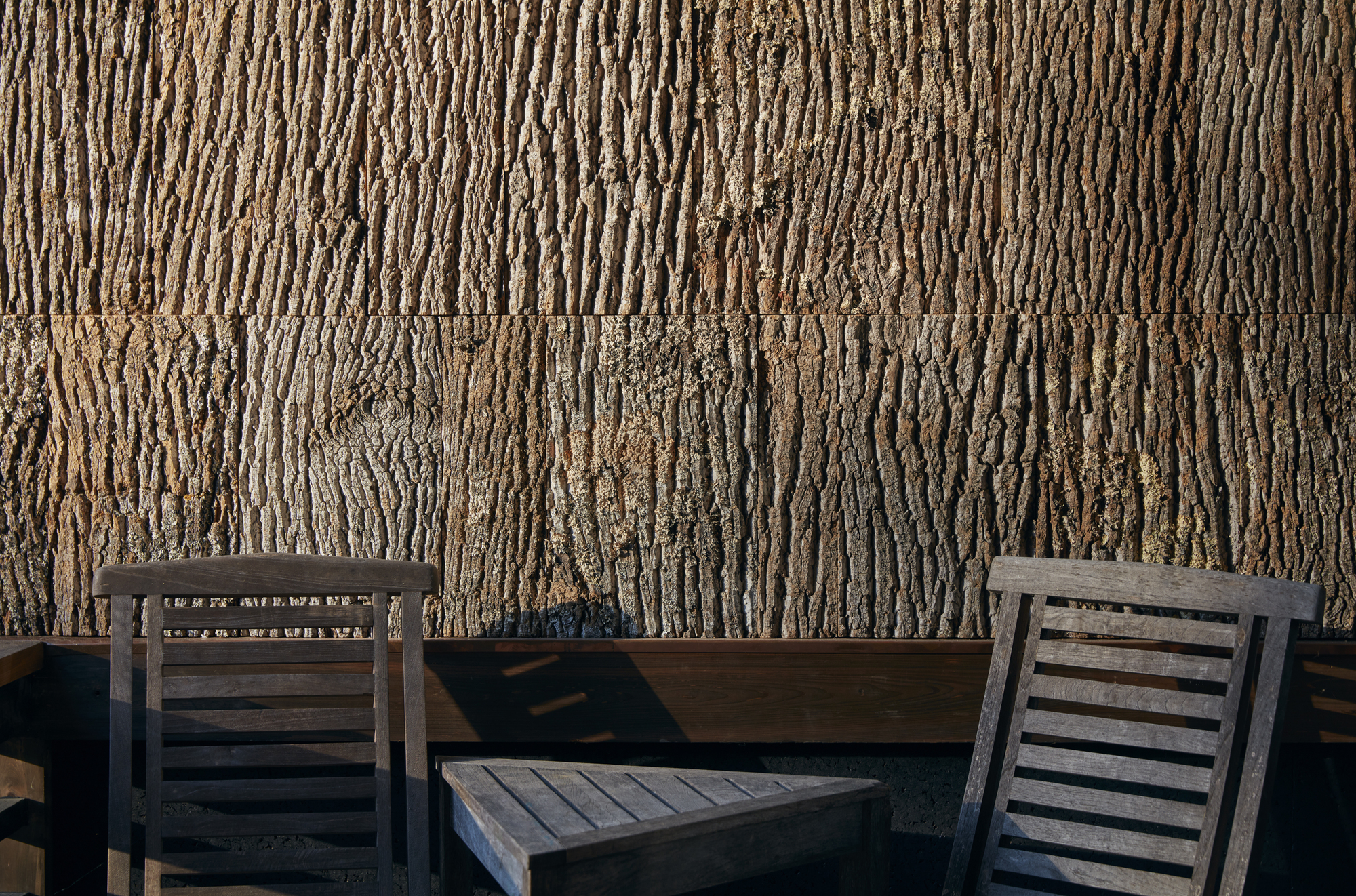 Detail of rough bark wall cladding with two timber chairs casting diagonal shadows on the deck