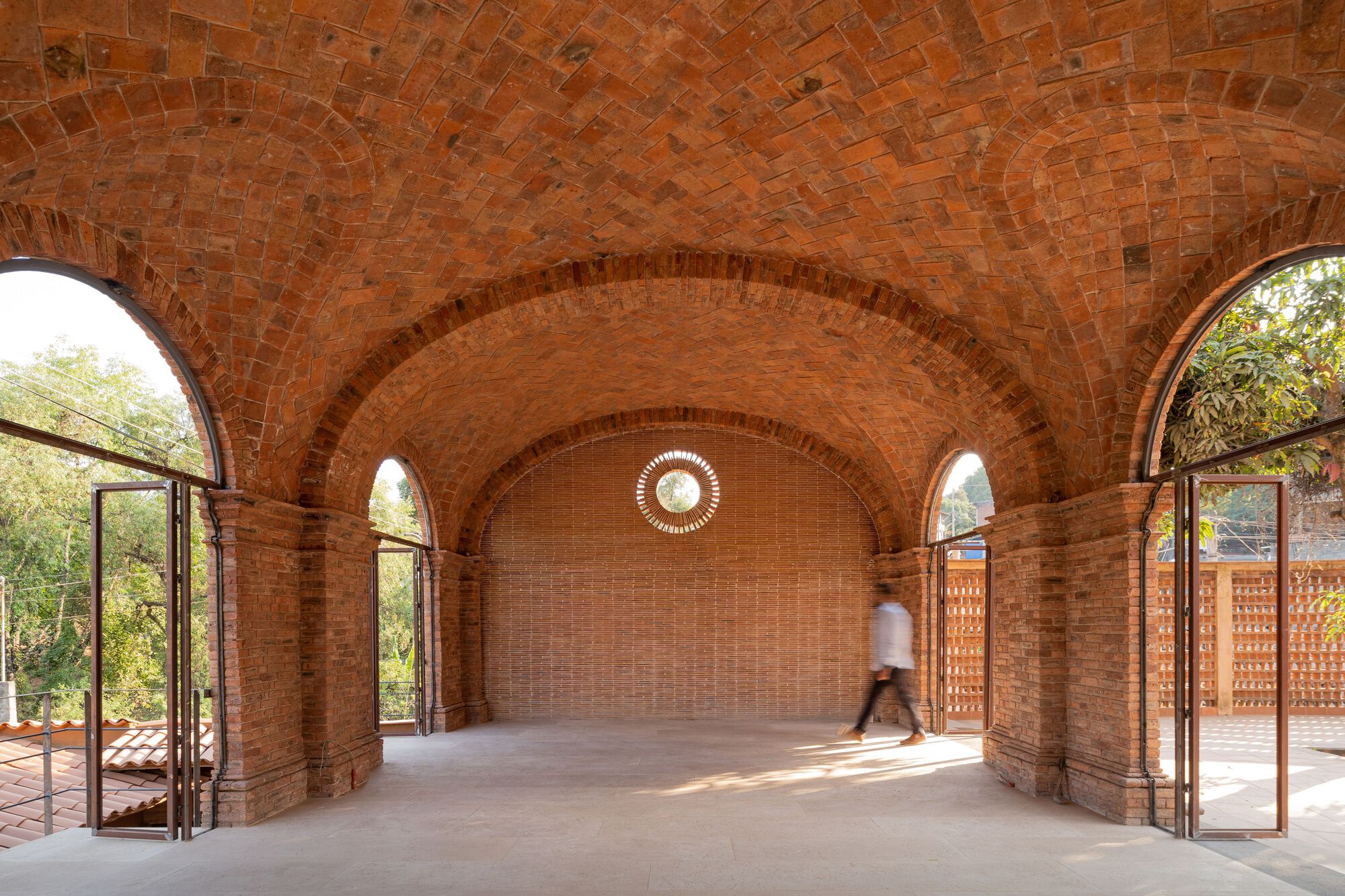 Interior of a brick vaulted hall with arched doorways and a figure walking through dappled sunlight