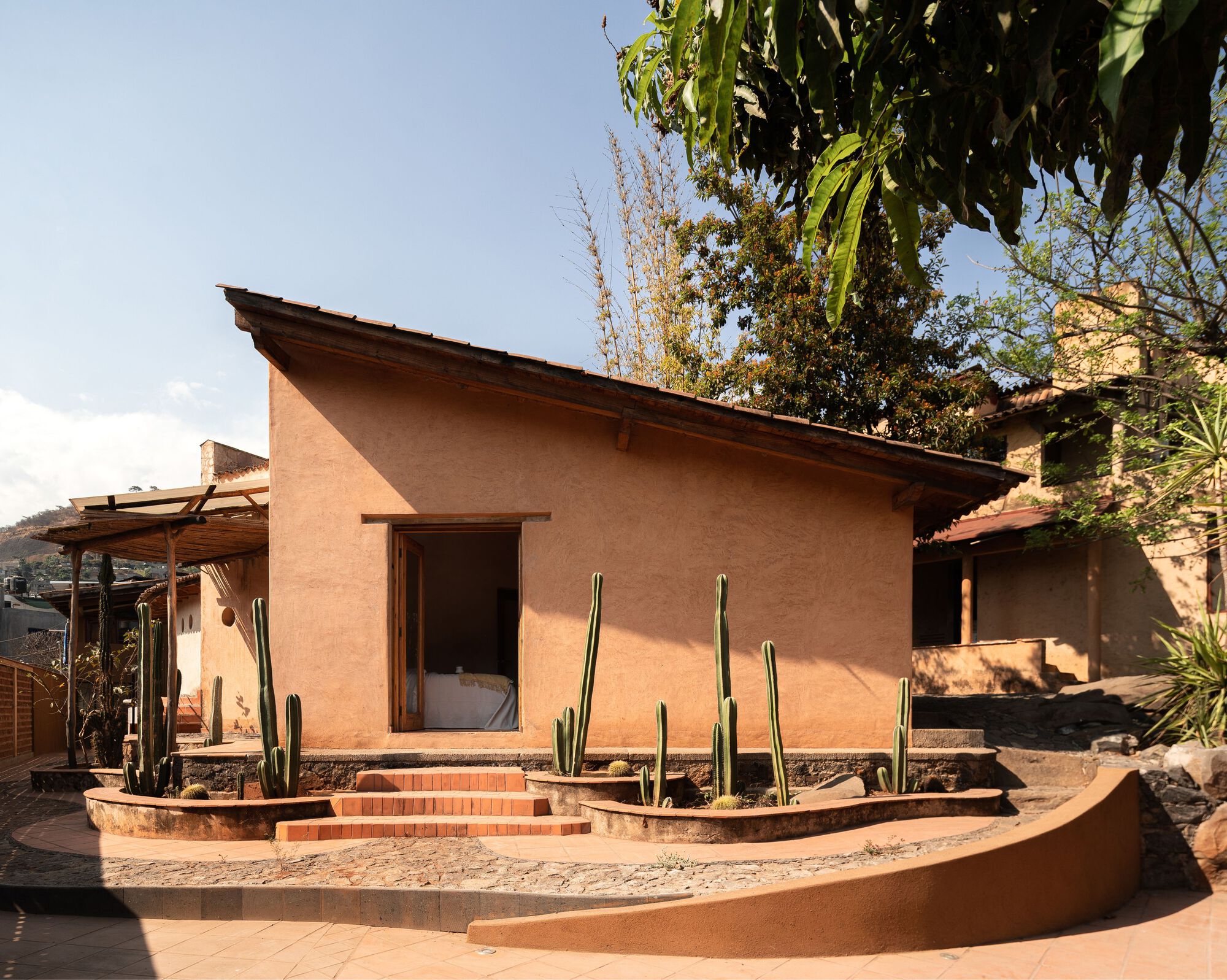 Earthen-plastered pavilion with angled roof beside terraced planting beds and cacti under afternoon light