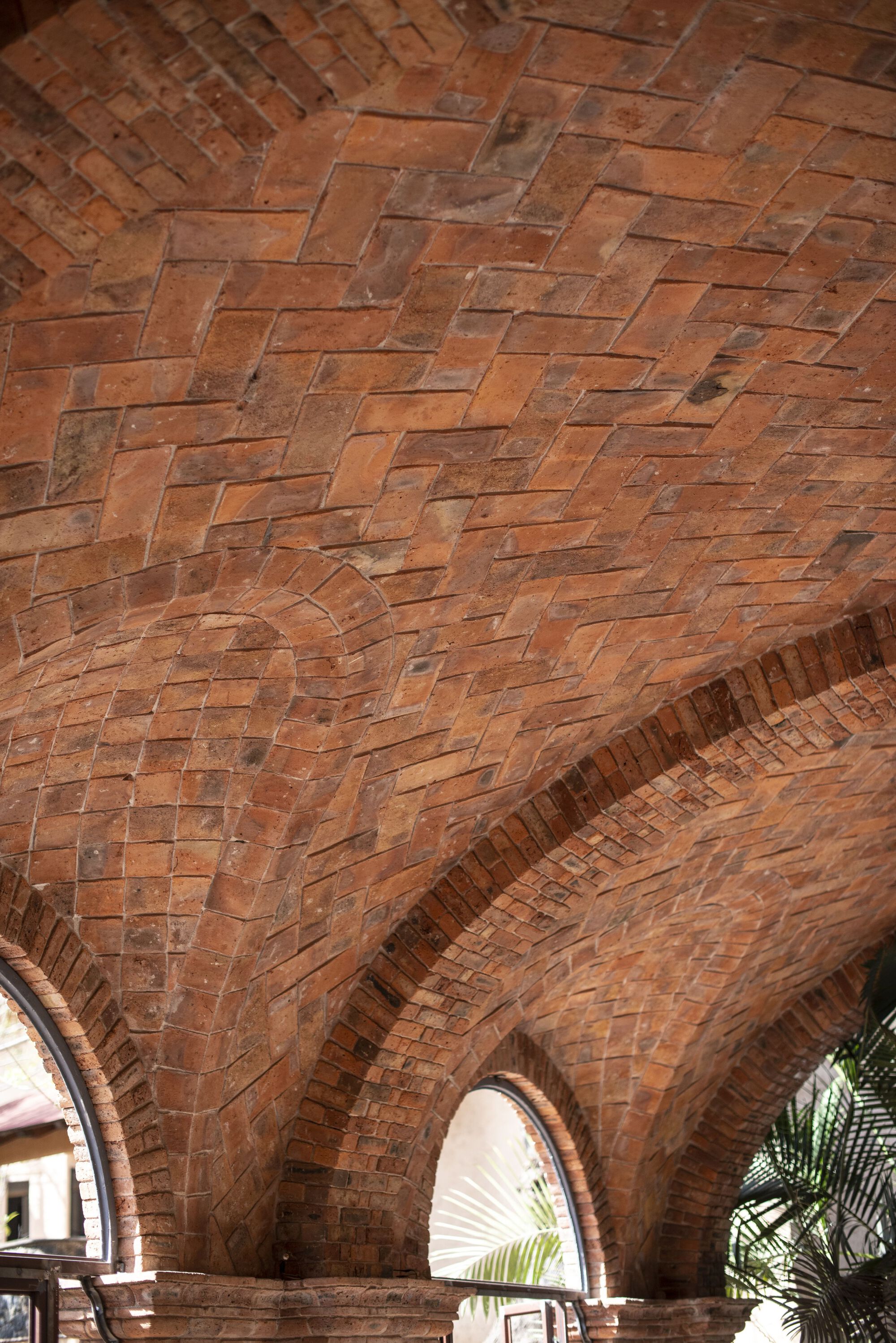 Vaulted brick ceiling in herringbone pattern with arched window openings framing palm fronds outside