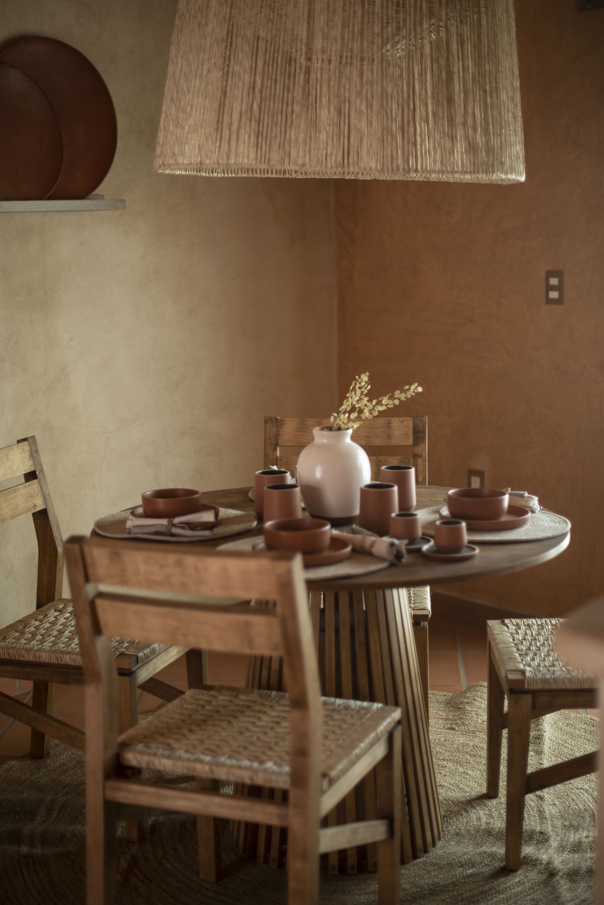 Round timber dining table with woven raffia pendant light and earthenware vessels in warm daylight