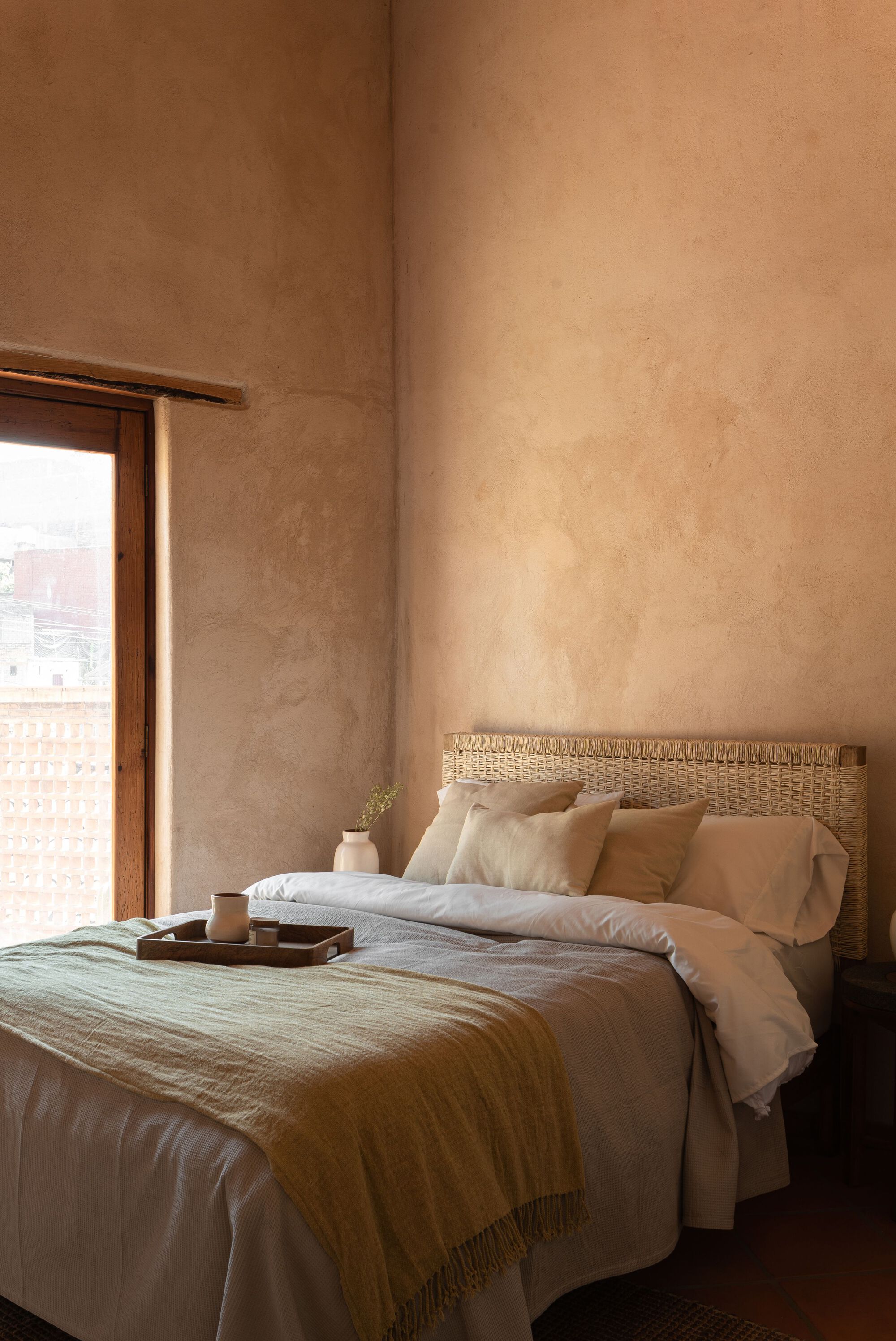 Bedroom corner with woven headboard and timber-framed window opening to distant rooftops