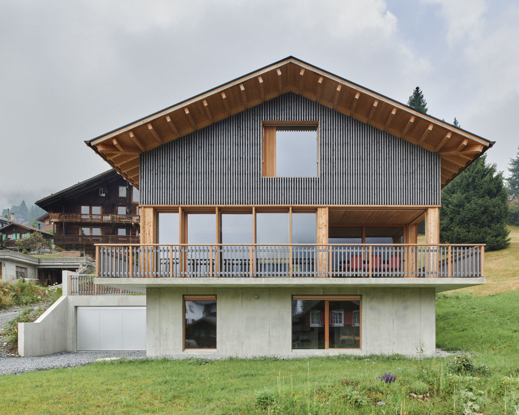 Gabled facade with vertical metal cladding and exposed timber eaves above a concrete base and grass slope