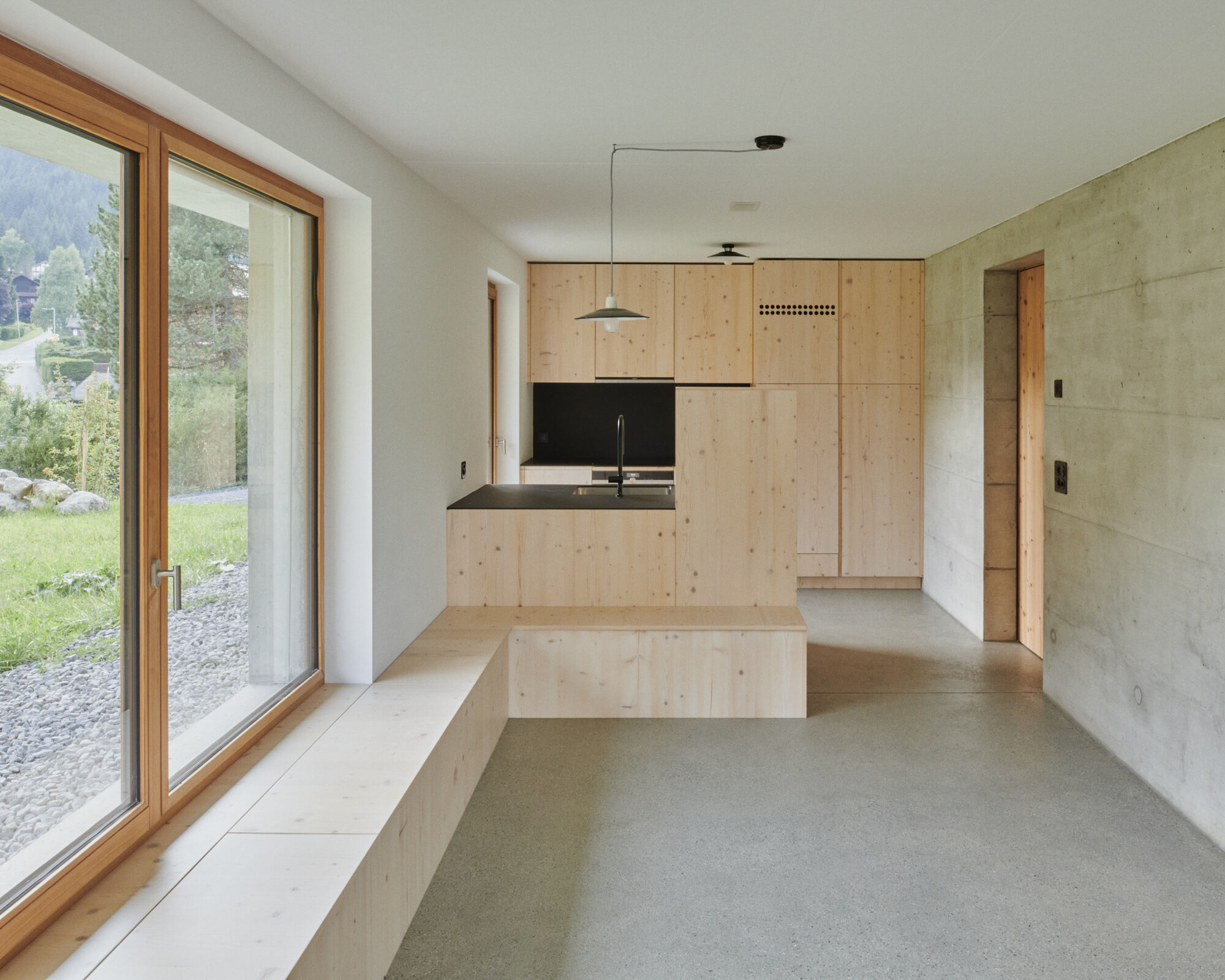 Kitchen with plywood cabinetry and black countertop alongside concrete walls and polished screed flooring