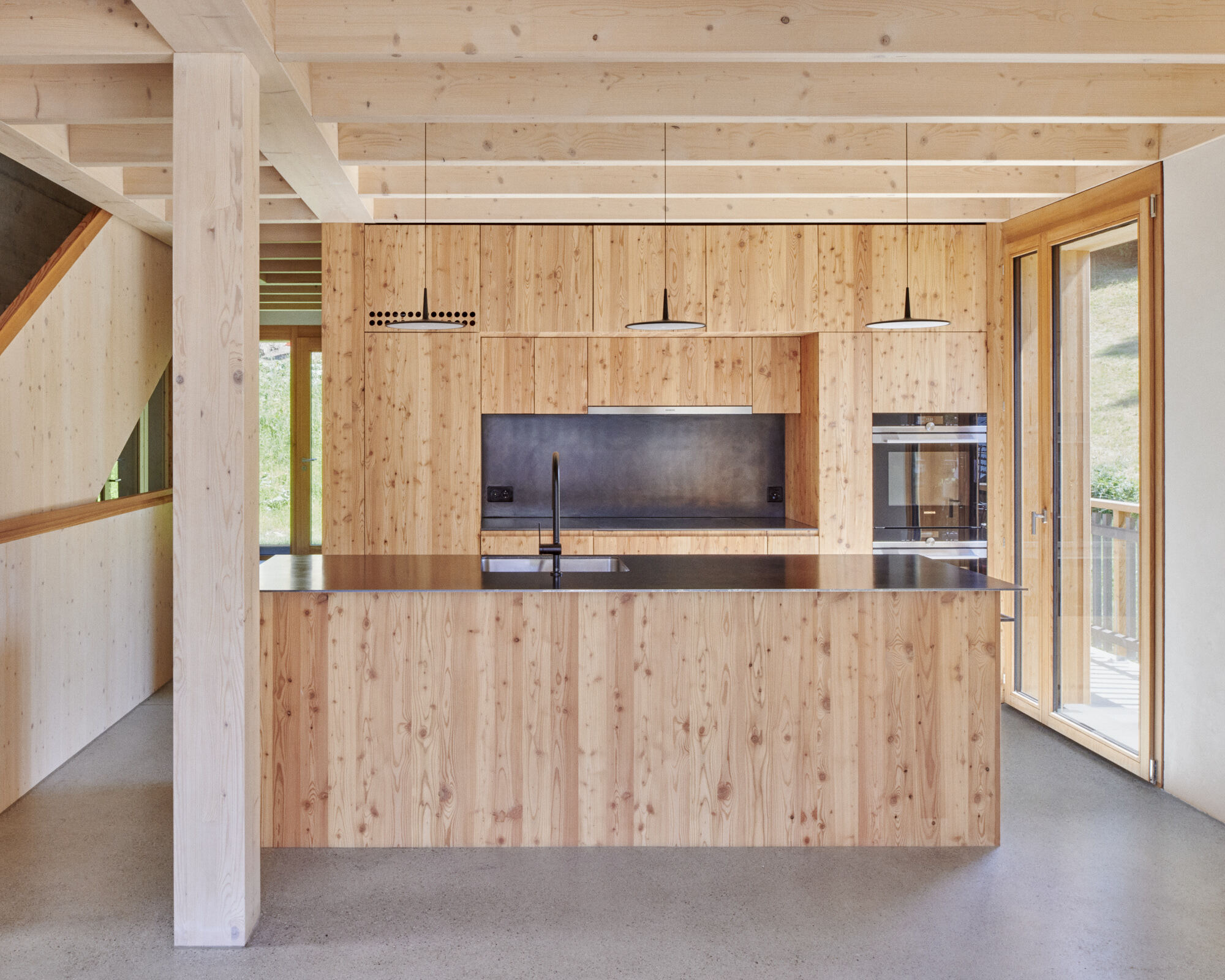 Open kitchen with vertical timber paneling and exposed ceiling beams overlooking green landscape through glazed doors