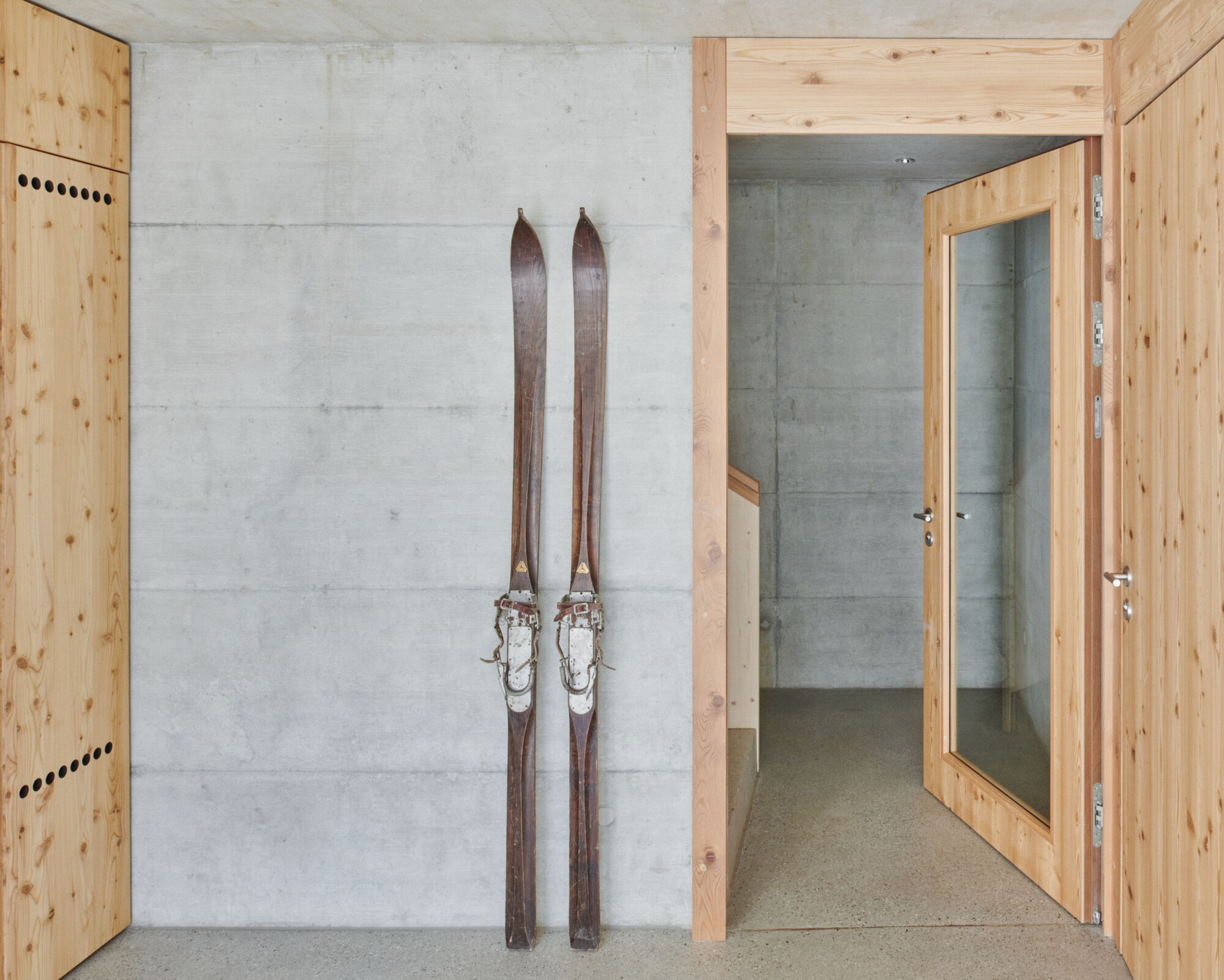 Pair of wooden skis leaning against a board-formed concrete wall beside timber-framed doorways