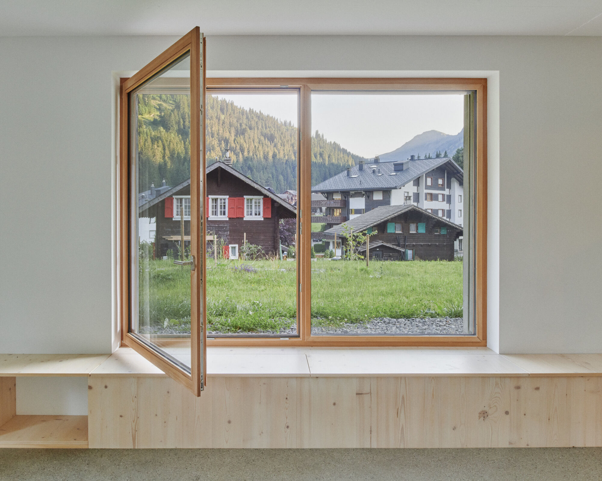 Interior view of timber-framed casement window opening to a meadow with village chalets and forested mountainside