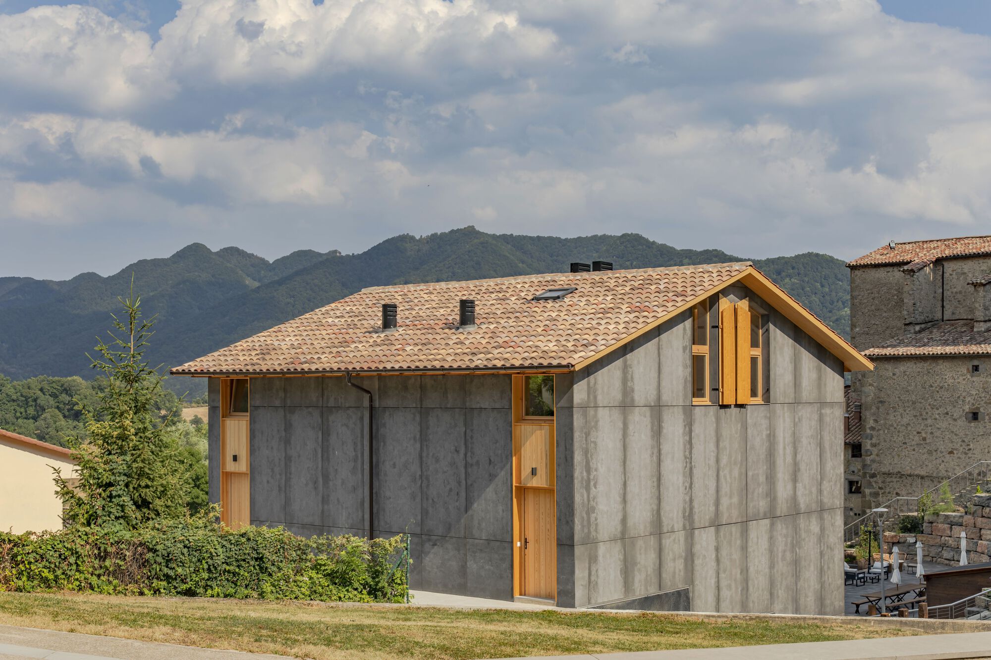 Tile-roofed structure with concrete panel walls and timber accents facing distant mountains under cloudy skies