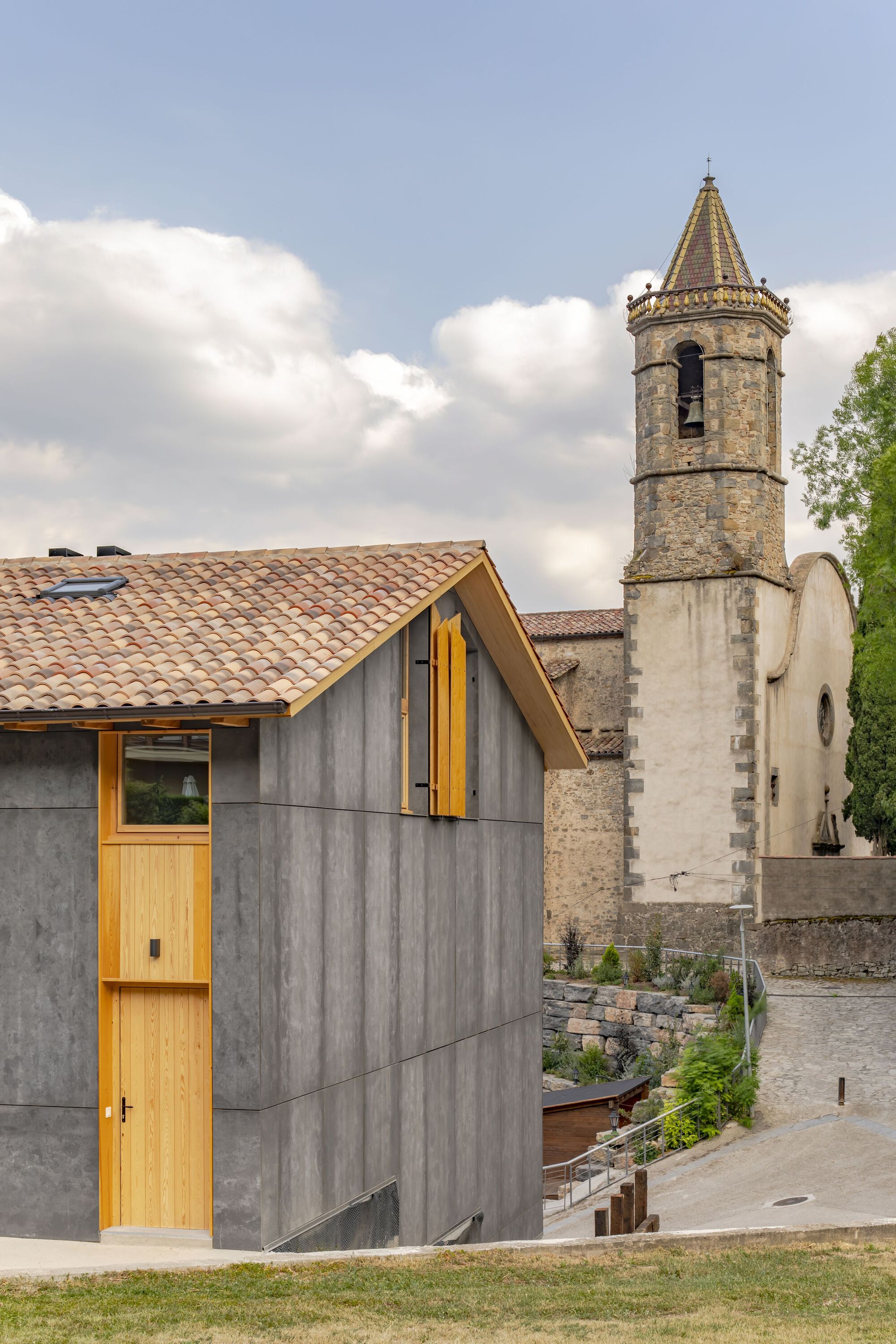 Board-formed concrete facade with timber-framed windows beside a stone bell tower under cloudy skies