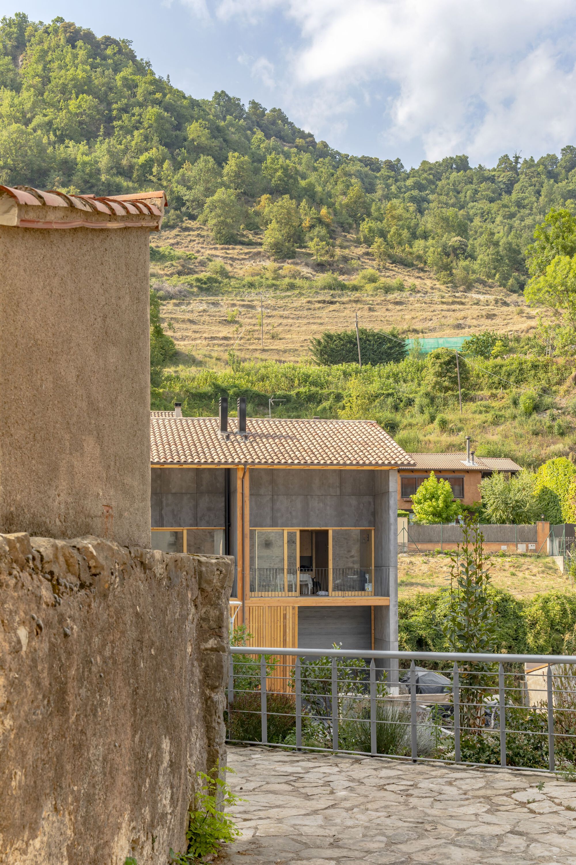 View of the terraced roof and concrete facade nestled among forested hills and surrounding village buildings