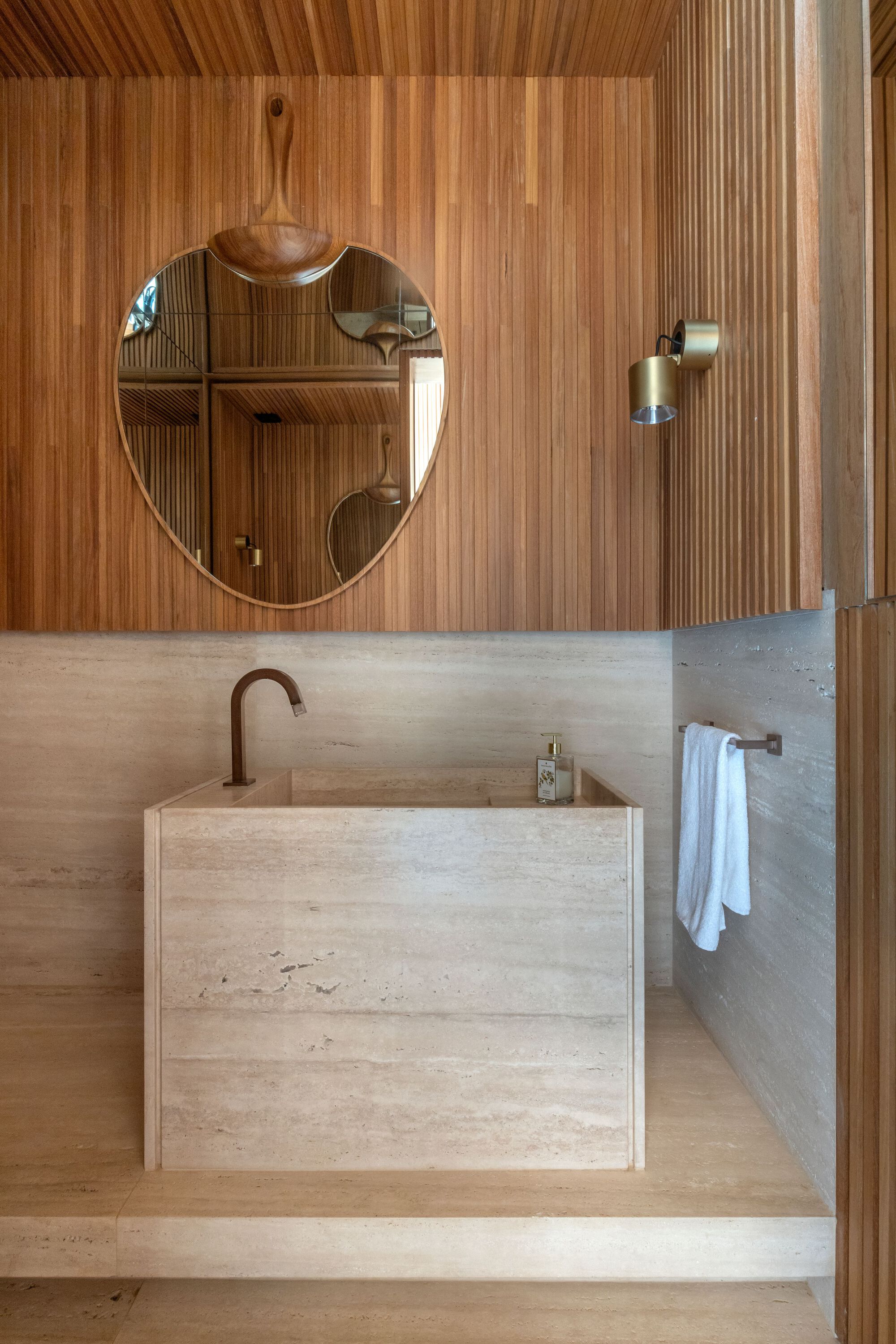 Bathroom vanity in travertine with bronze fixtures beneath ribbed timber wall cladding and round mirror