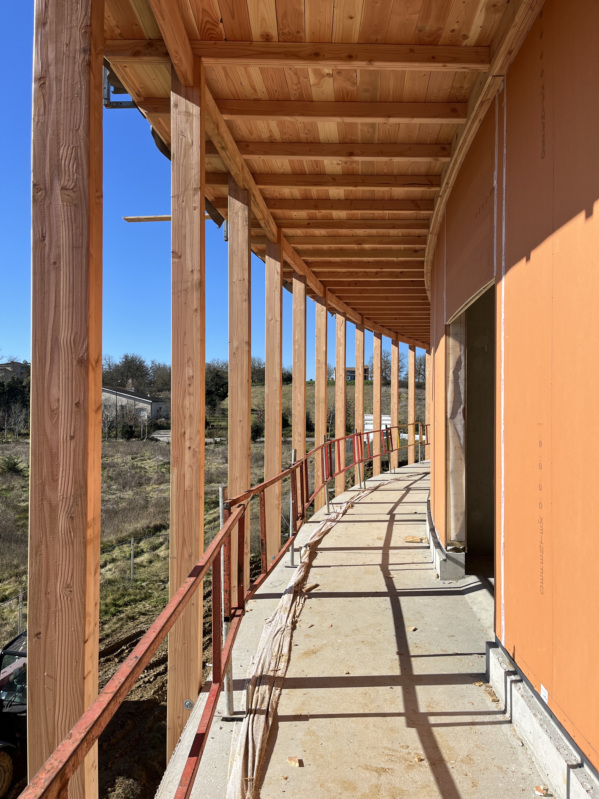 Exterior walkway with exposed timber beams, red metal railing and concrete floor casting afternoon shadows