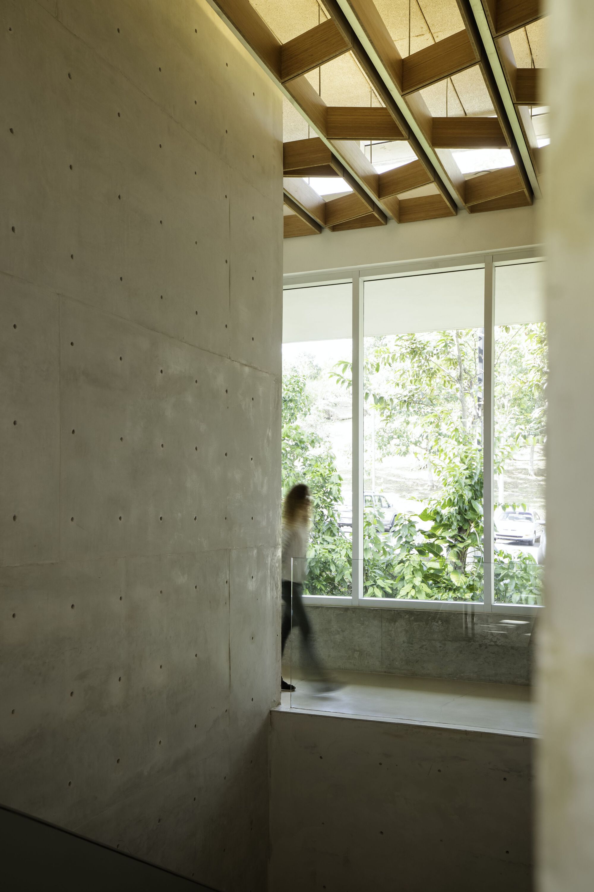 Interior view of concrete wall beside tall window with blurred figure passing through skylit timber-beamed space