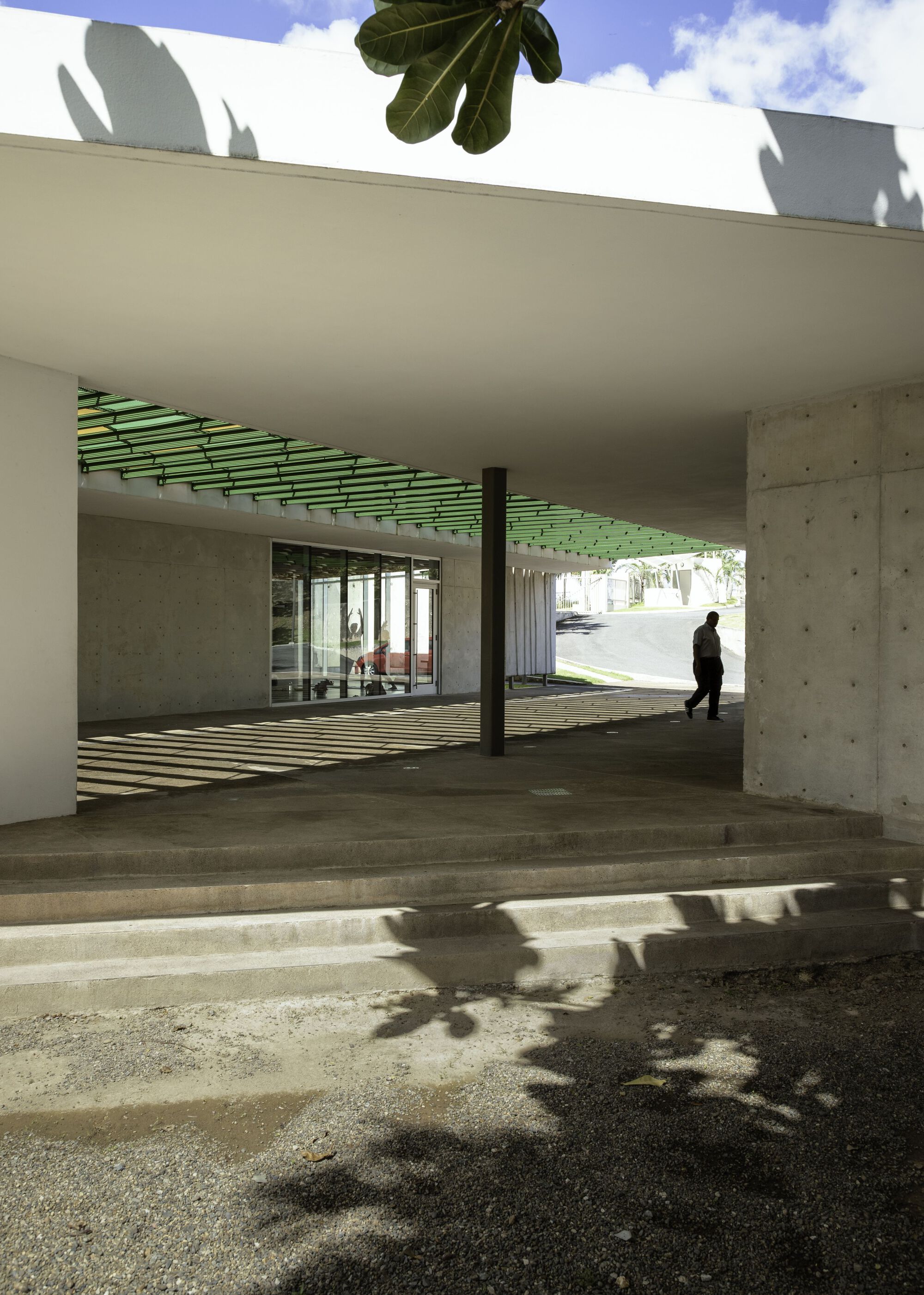 Covered concrete terrace with green slatted ceiling, shadow patterns on floor, and single figure walking through