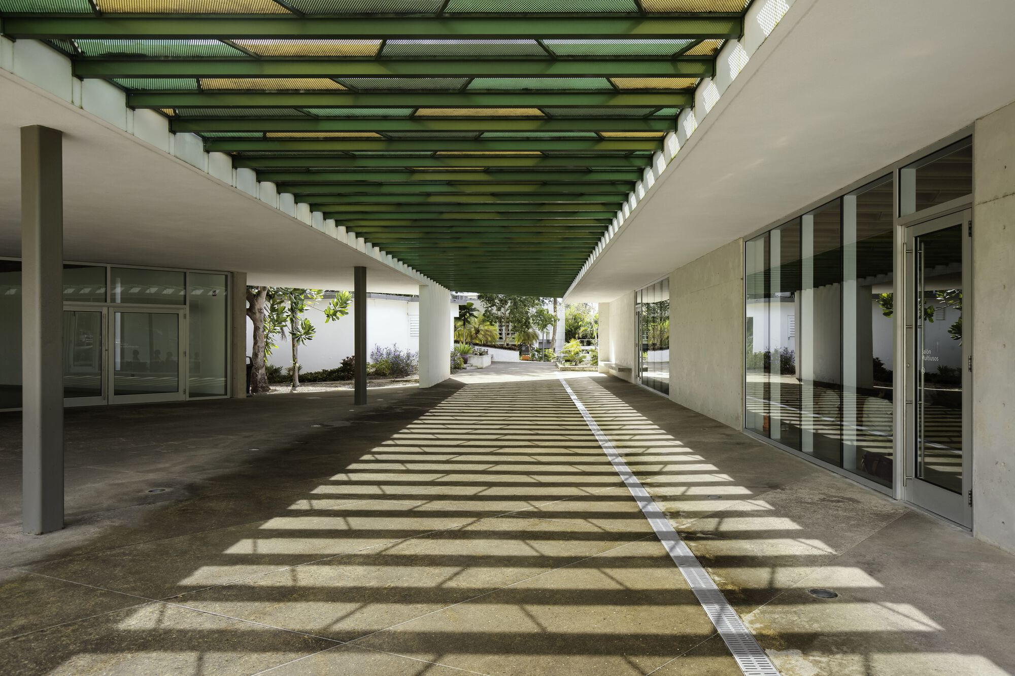 Covered walkway with green corrugated ceiling panels casting striped shadows on concrete pavement