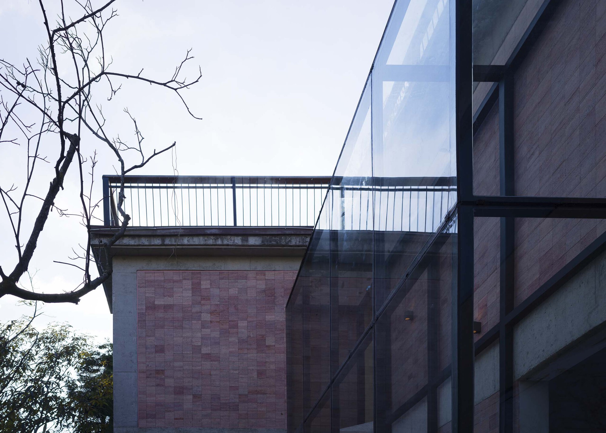 Glass facade reflecting rammed earth tile wall and bare tree branches under an overcast sky