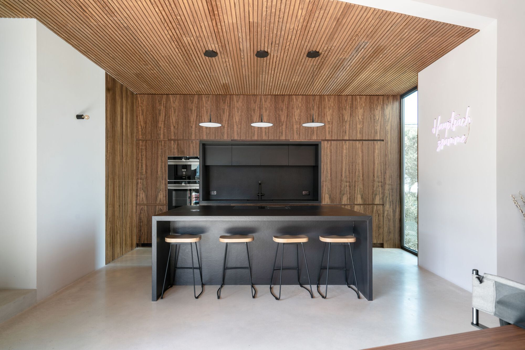 Kitchen island with four stools below timber slat ceiling and walnut cabinetry wall with pendant lights