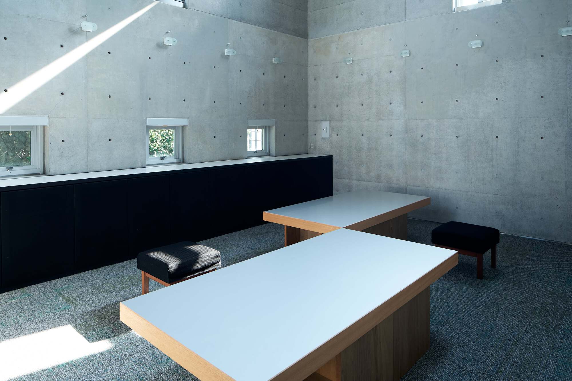 Board-formed concrete interior with low tables and cushioned stools on carpet under sloped skylights