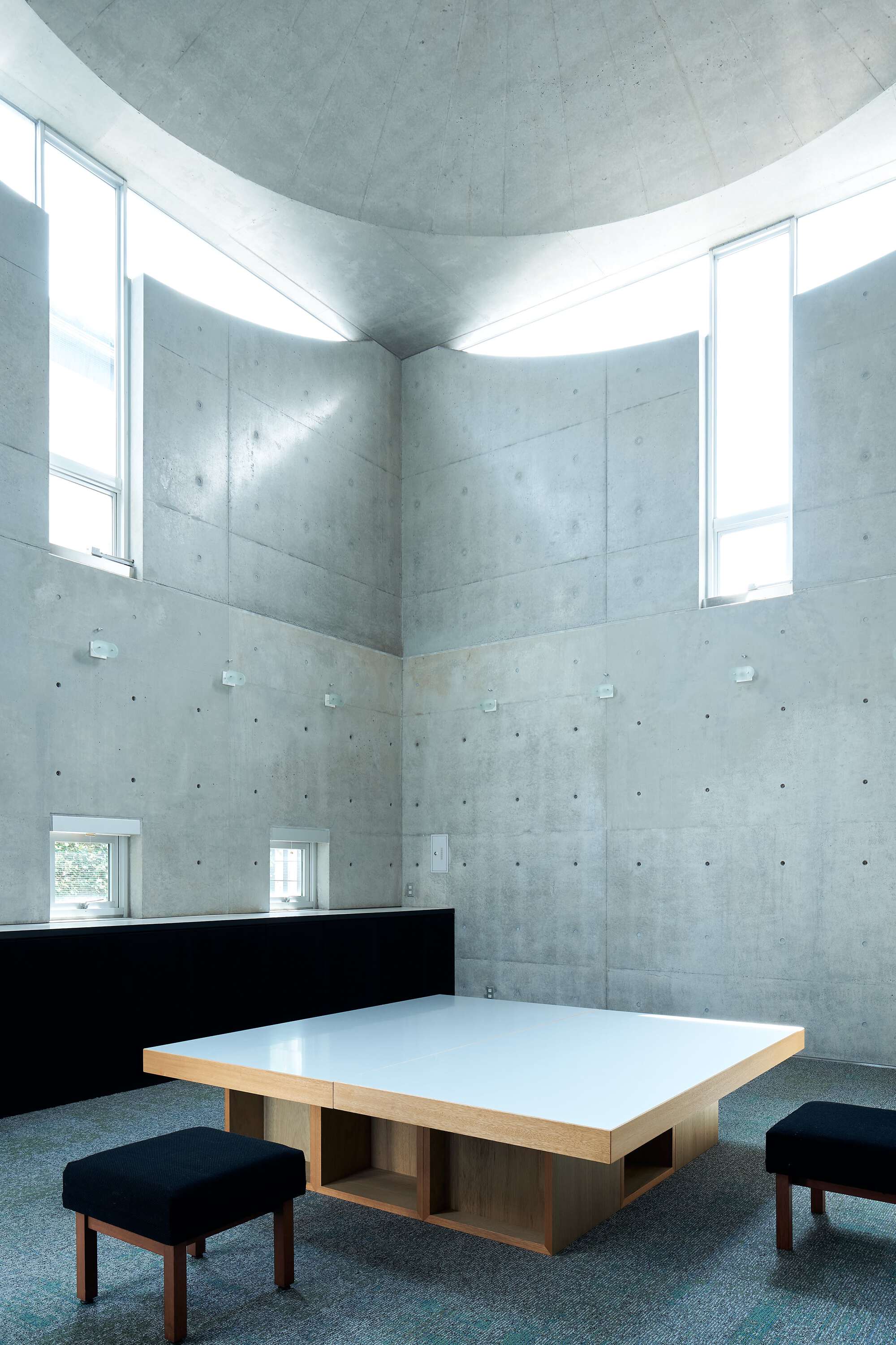 Vaulted concrete space with clerestory windows illuminating walls and low table with stools below
