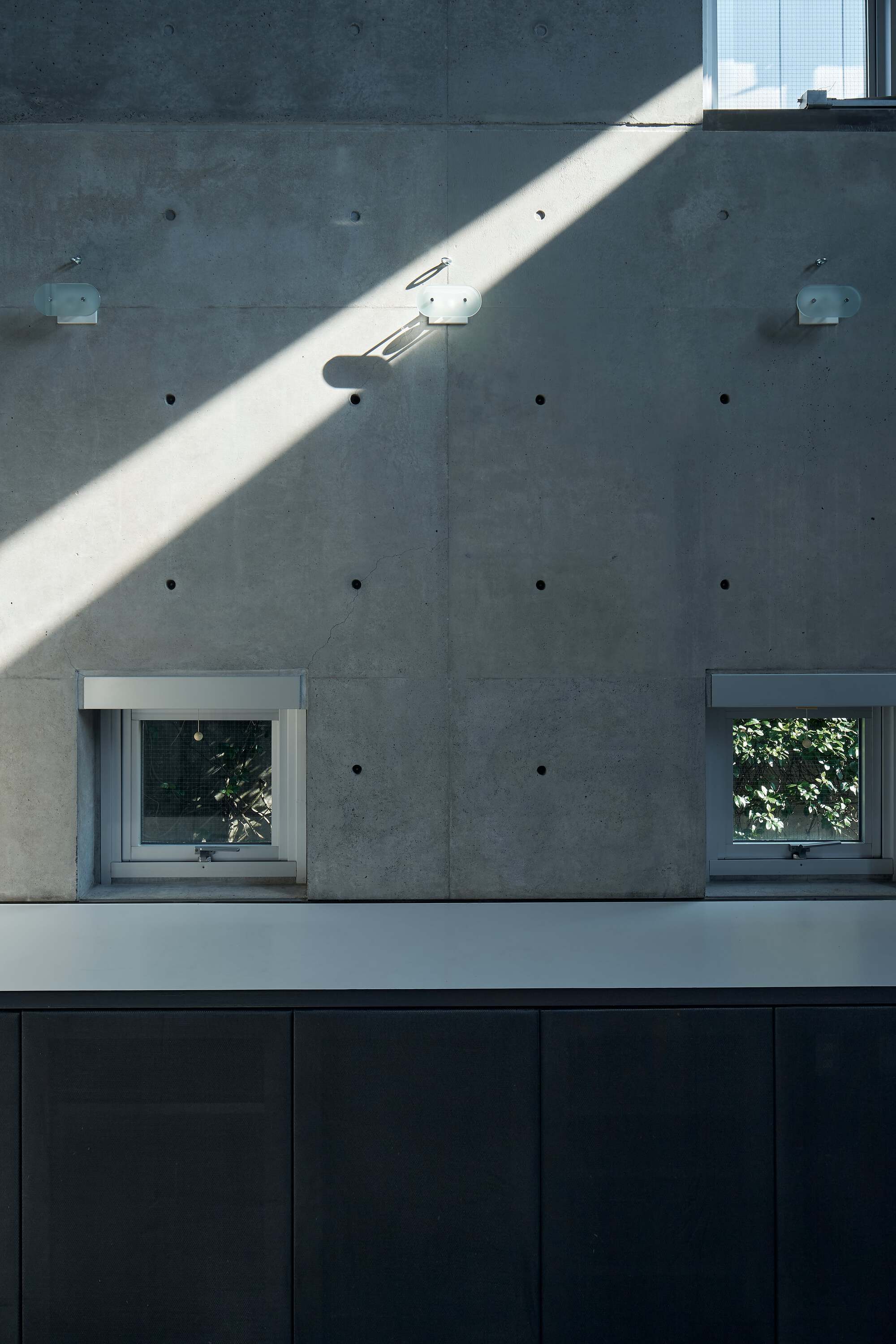Board-formed concrete wall with two square windows and a diagonal shaft of afternoon sunlight above dark cabinetry