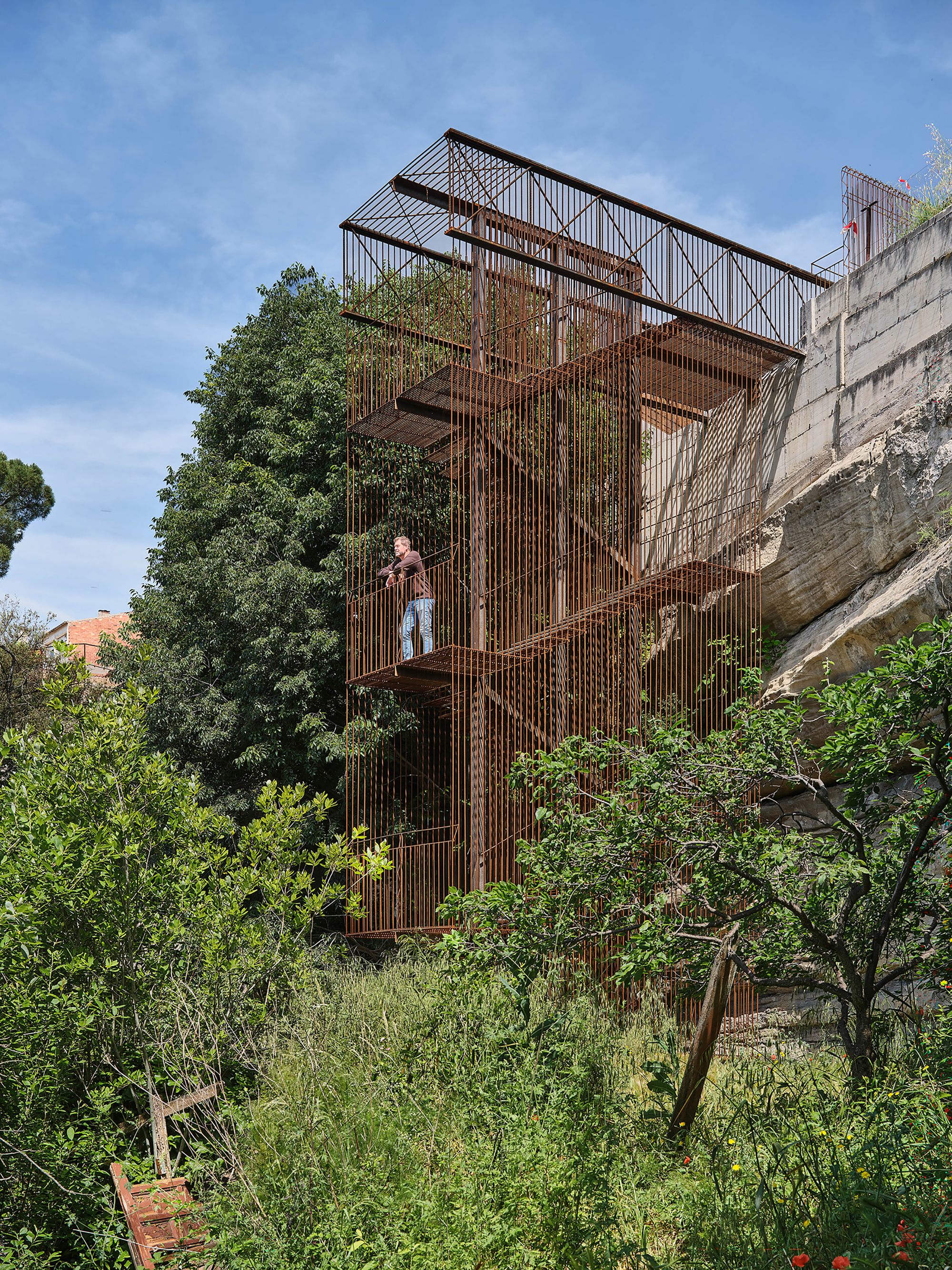 Vertical corten steel staircase tower with a person standing on a platform surrounded by dense vegetation
