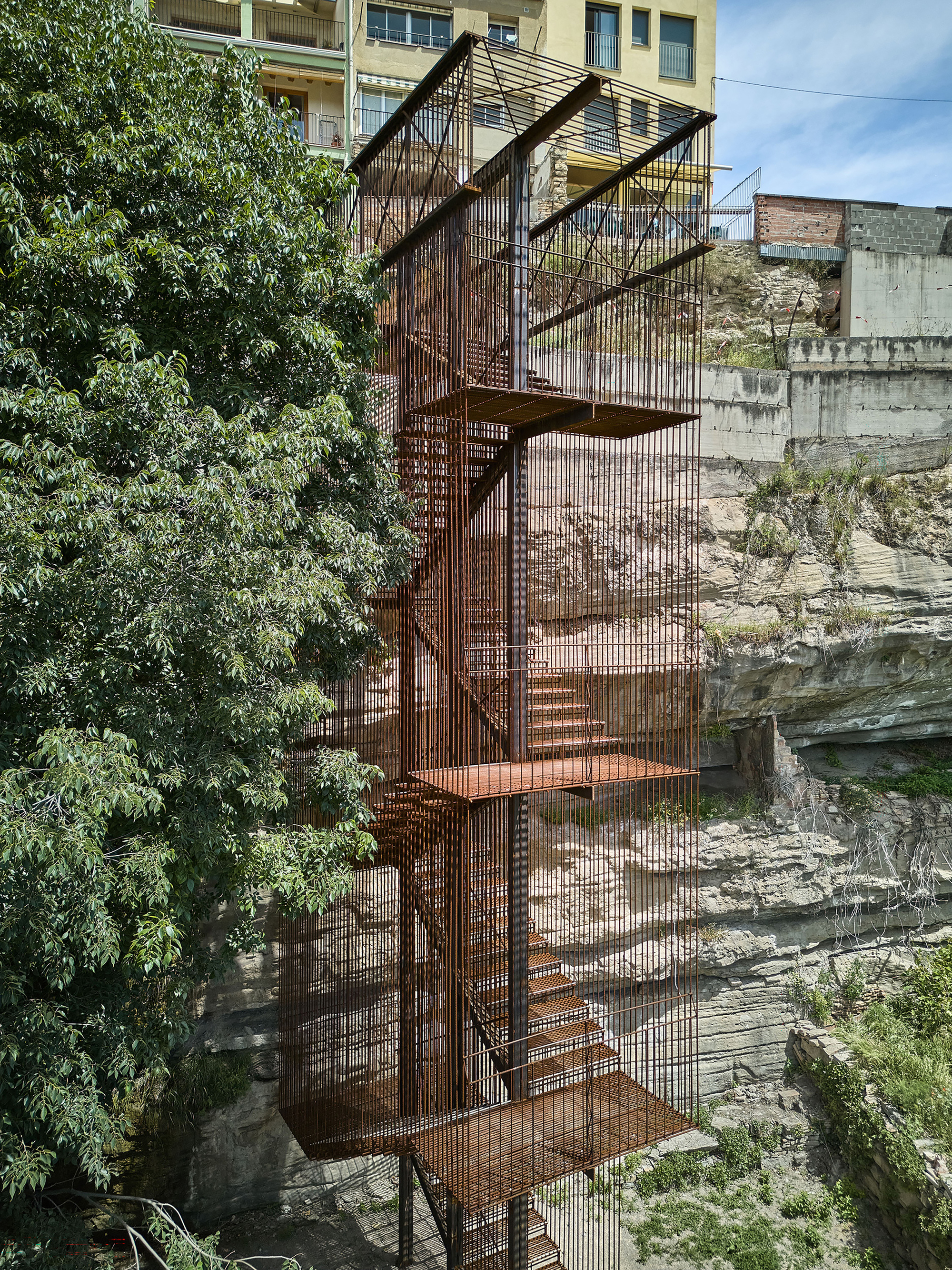 Frontal view of the perforated steel stair tower emerging from layered sedimentary rock beside mature trees