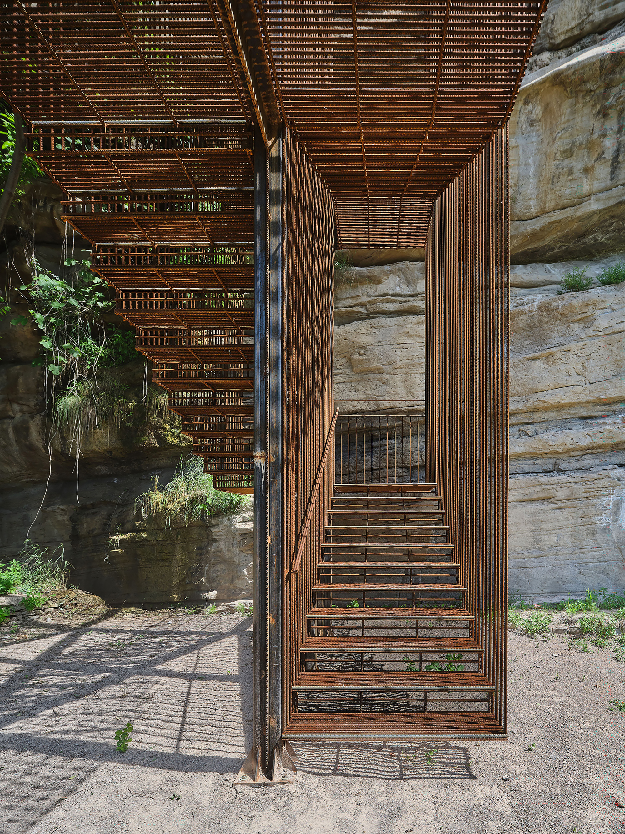 Looking up through the staircase core with weathered steel slats and exposed rock walls casting shadows