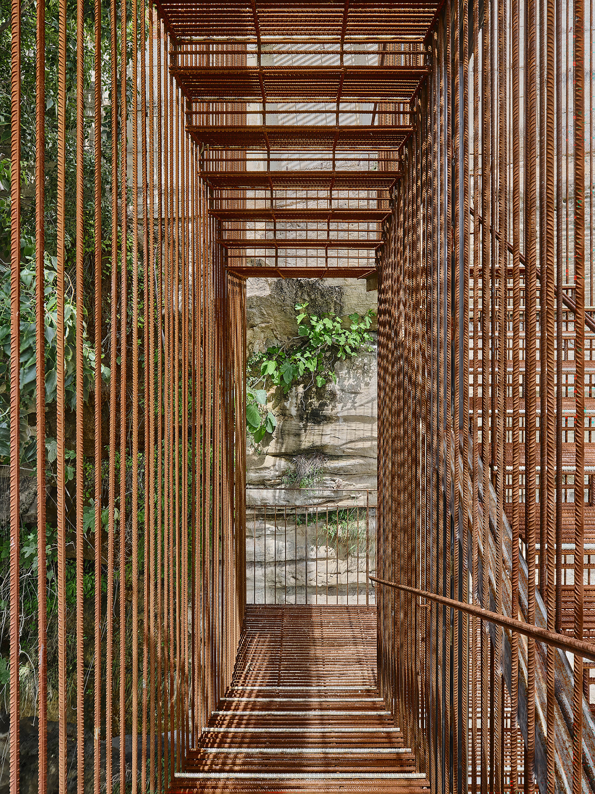 View through the narrow metal stair enclosure with rod balusters framing weathered stone and vegetation below