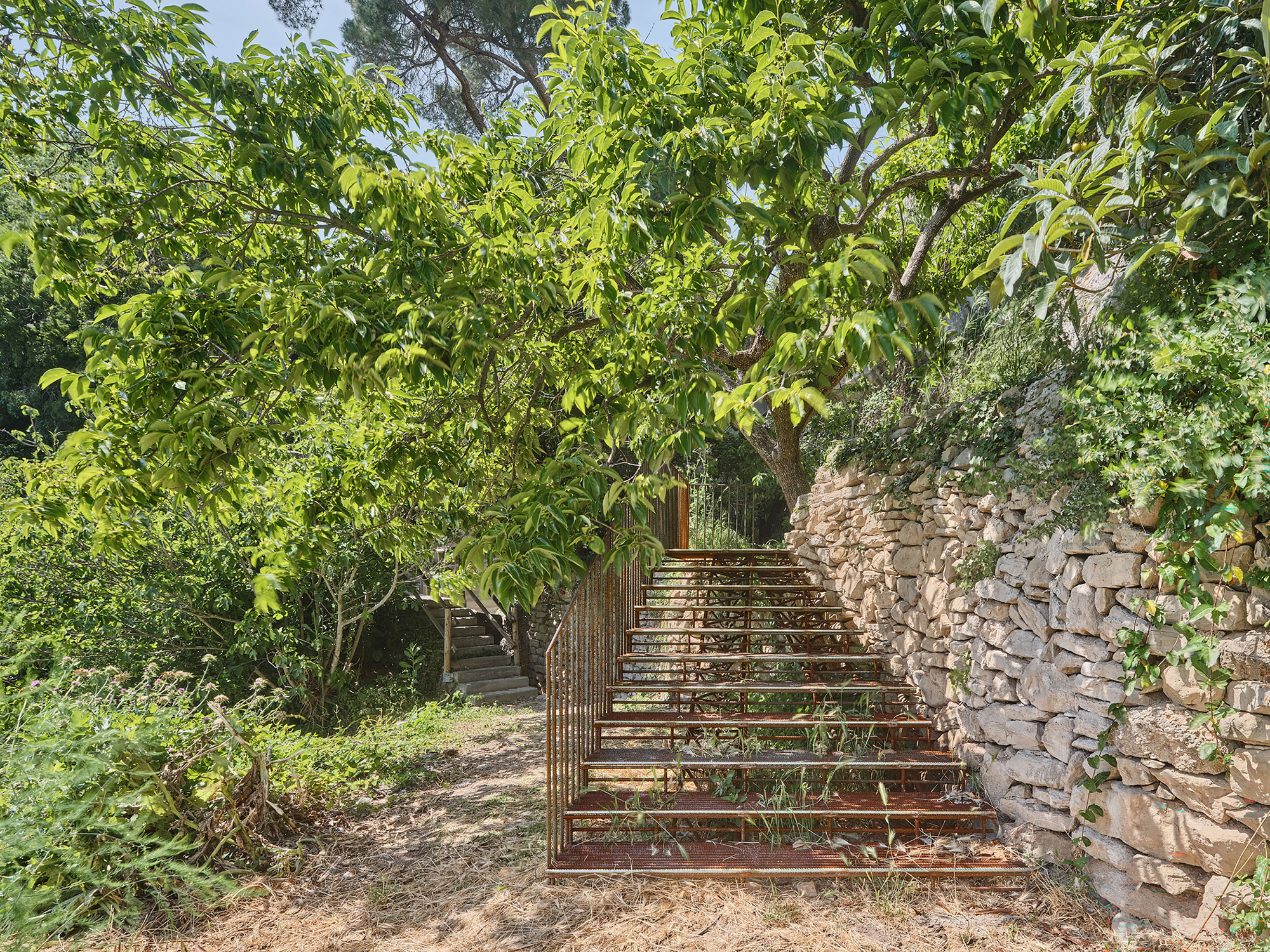 Metal frame stair structure under leafy tree canopy beside a dry-stacked stone retaining wall