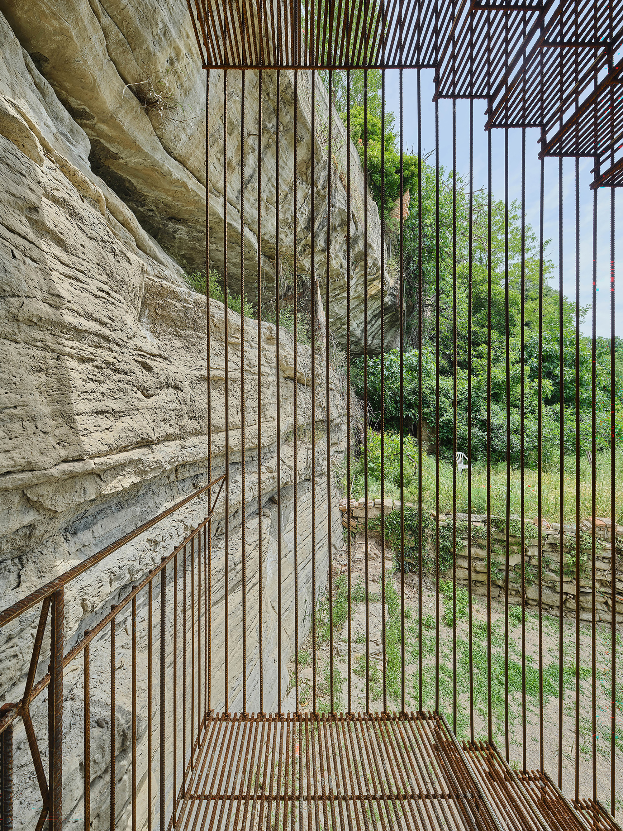Interior view through vertical rod balusters with stone cliff face on one side and green landscape beyond