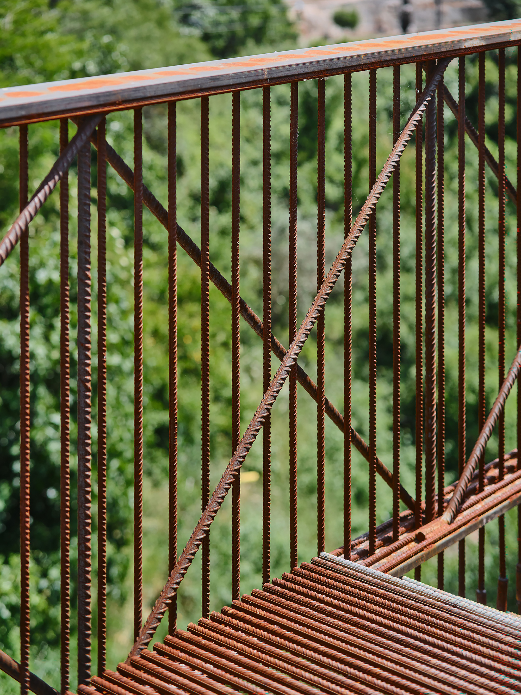 Exposed rebar railing with diagonal cross-bracing against a backdrop of green foliage