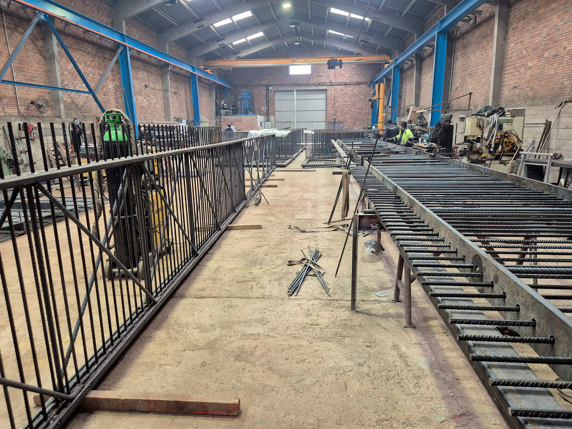 Fabrication workshop with rebar assemblies and a worker in high-visibility vest under blue steel trusses