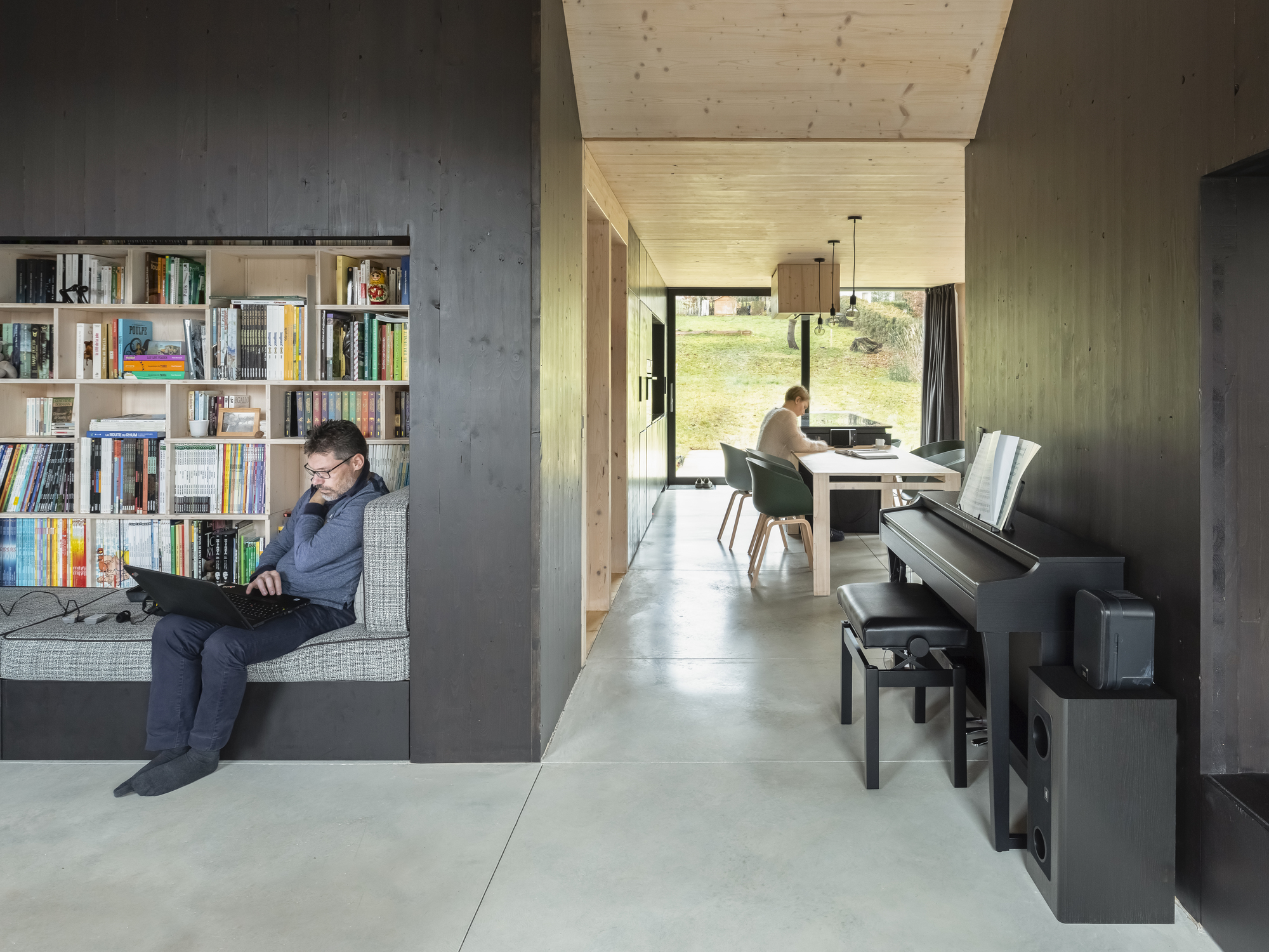 Interior view through concrete walls showing a man reading by the bookshelf and a woman at the desk