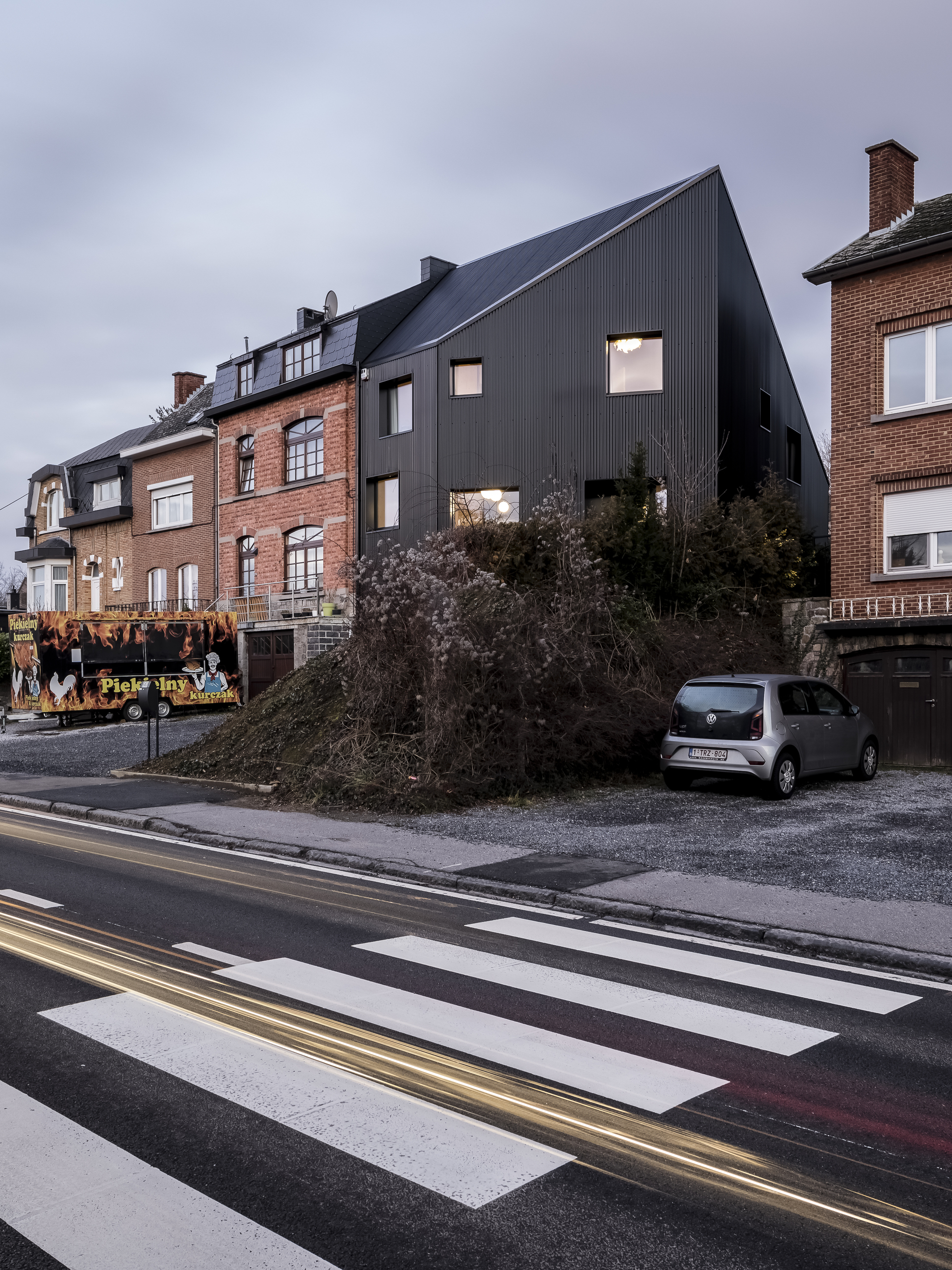 Street view of the dark corrugated metal facade rising above neighboring brick houses under an overcast sky