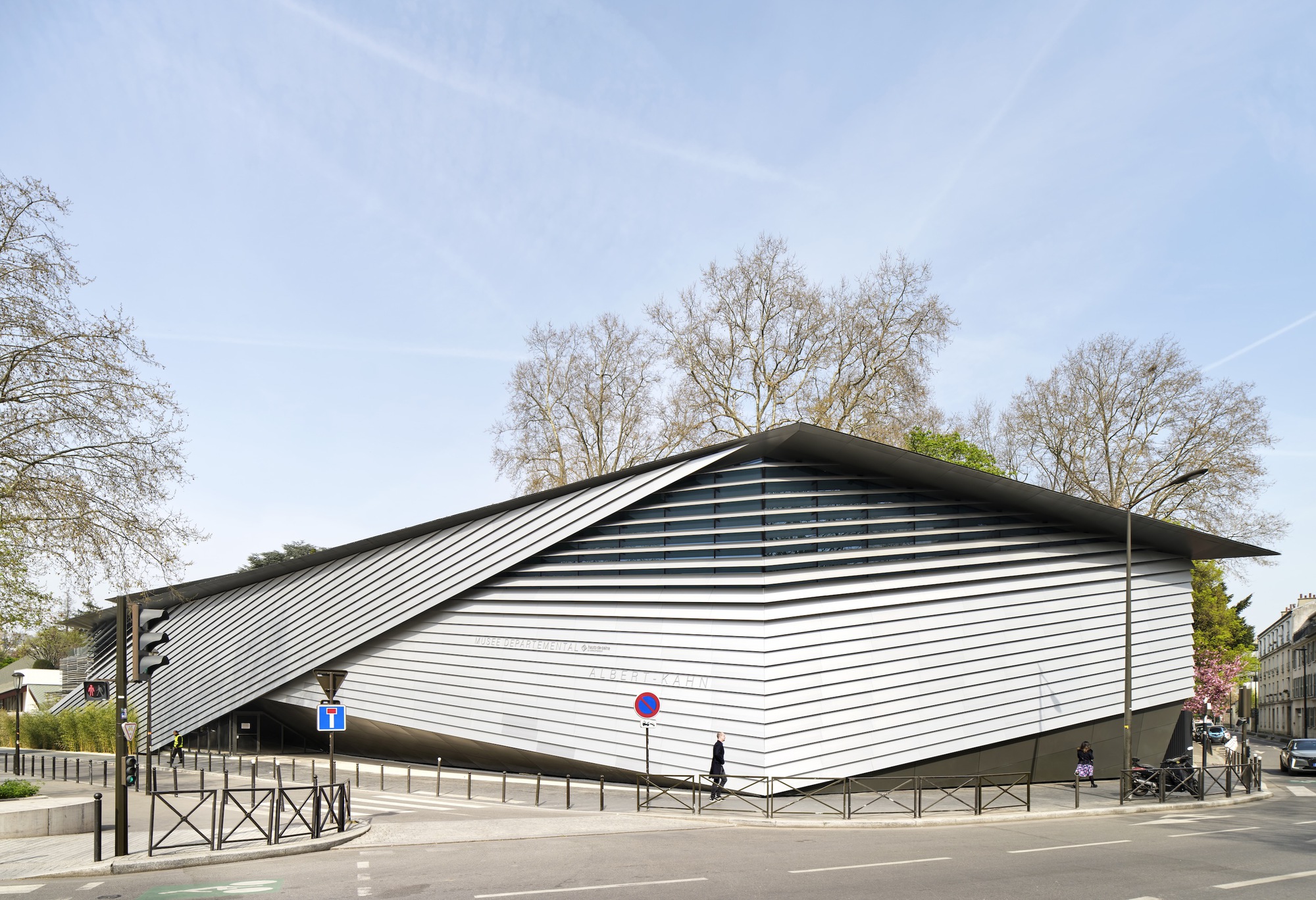 Street view of the sculptural ribbed metal facade with bare trees in the background