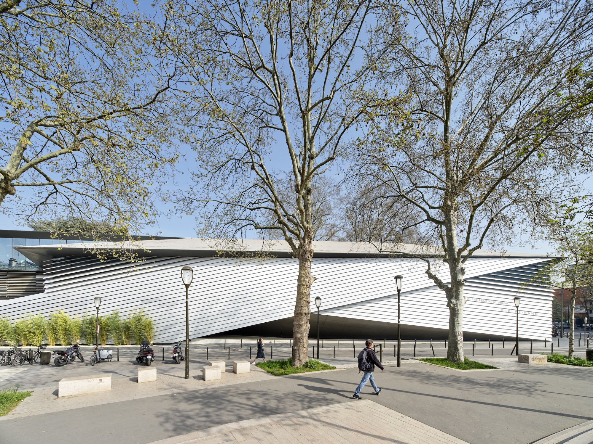 Horizontal metal louvers wrapping the curved facade as pedestrians walk beneath spring trees