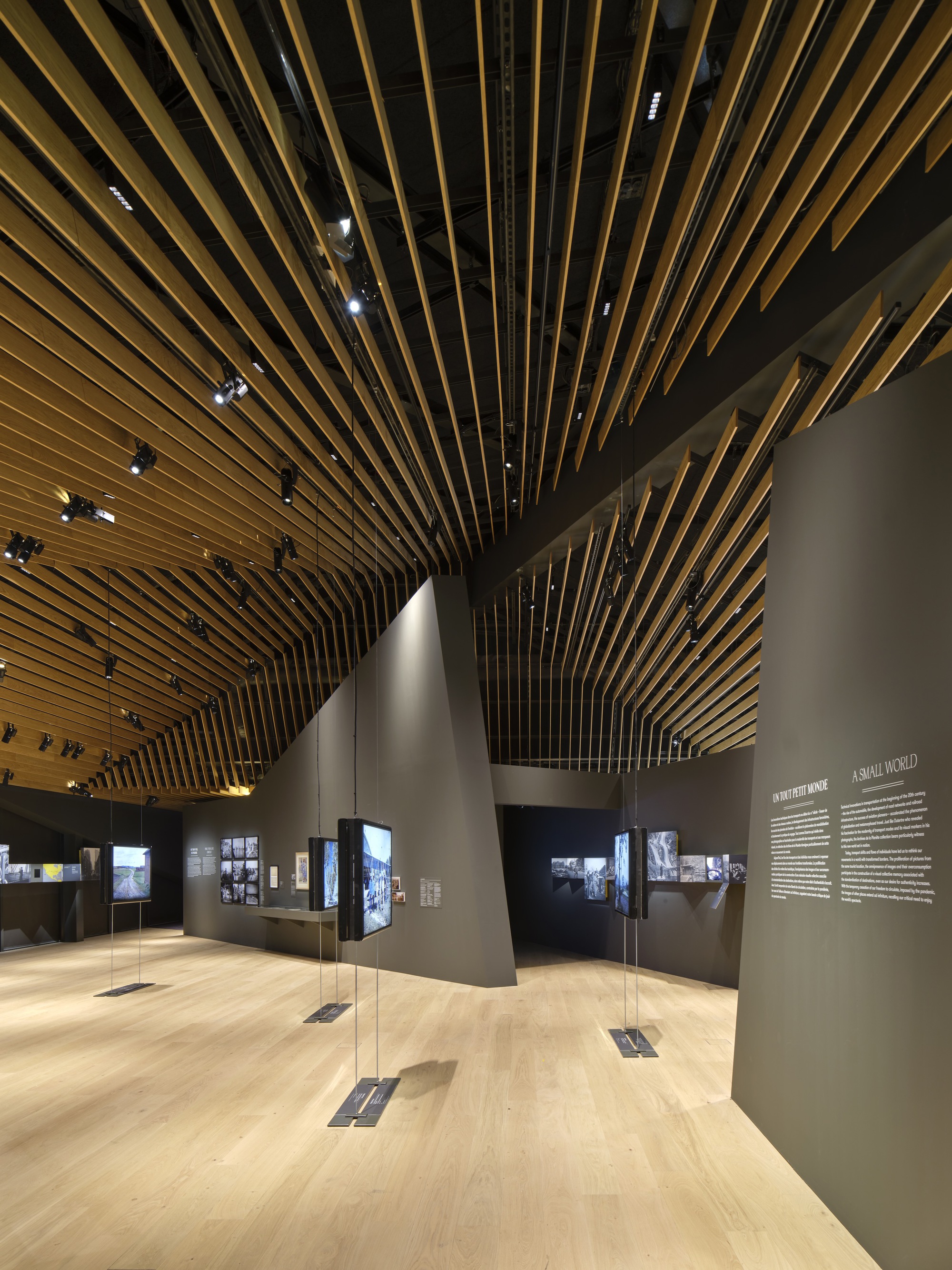 Exhibition gallery with radiating timber baffle ceiling and grey partition walls displaying photographs and text panels