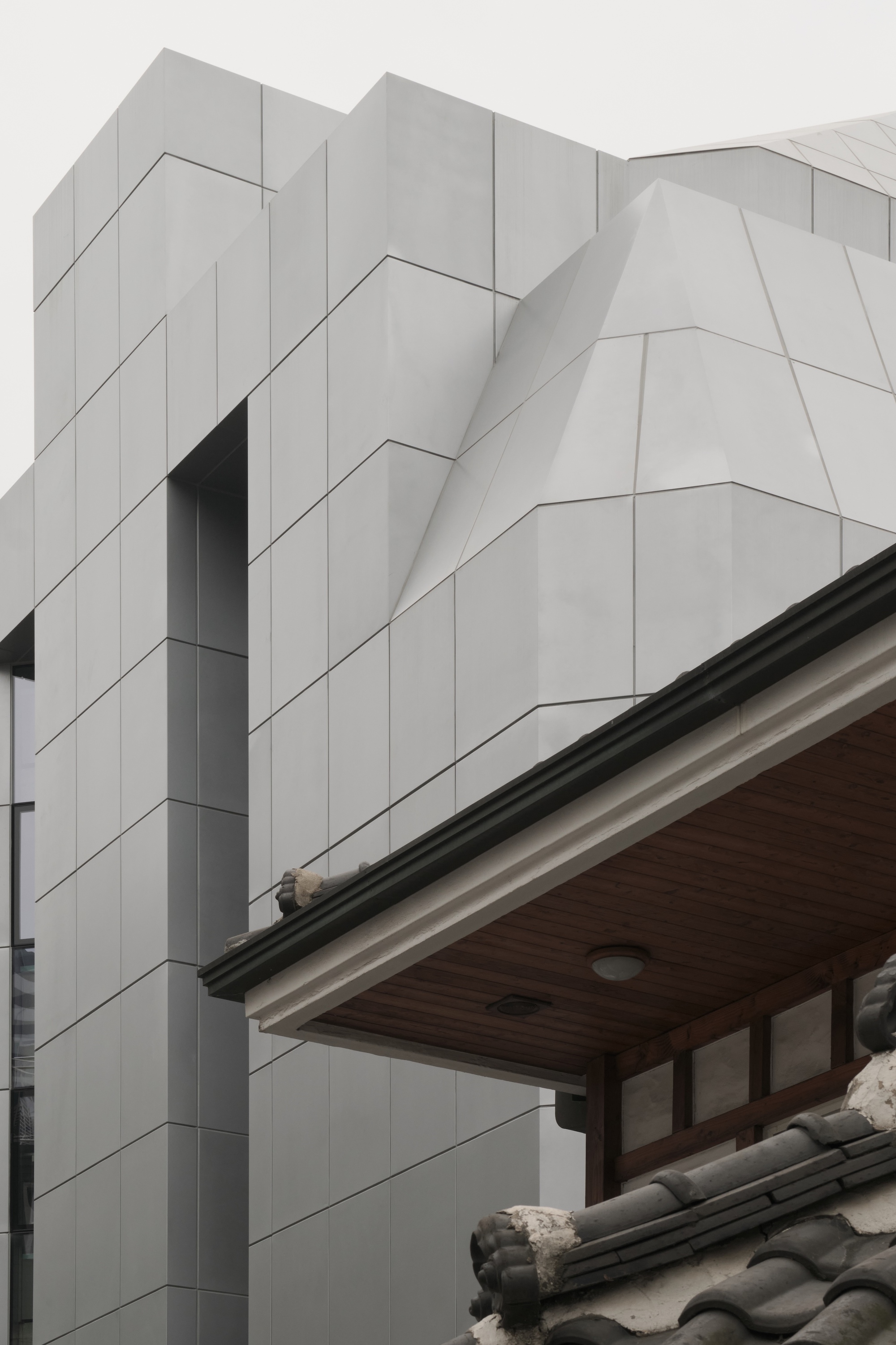 Faceted white panel cladding rising beside a tiled traditional roof under cloudy skies