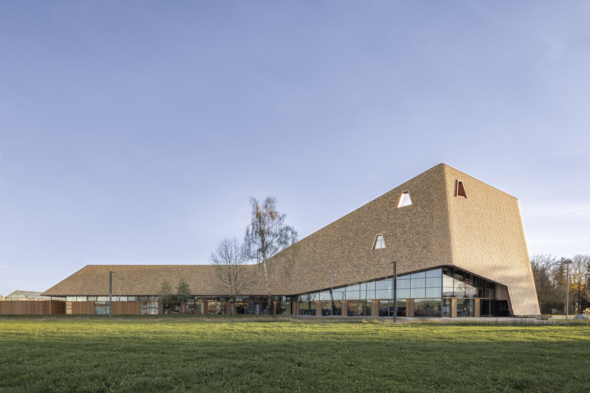 Side view of the shingled facade with triangular openings and glazed base across a lawn at dusk