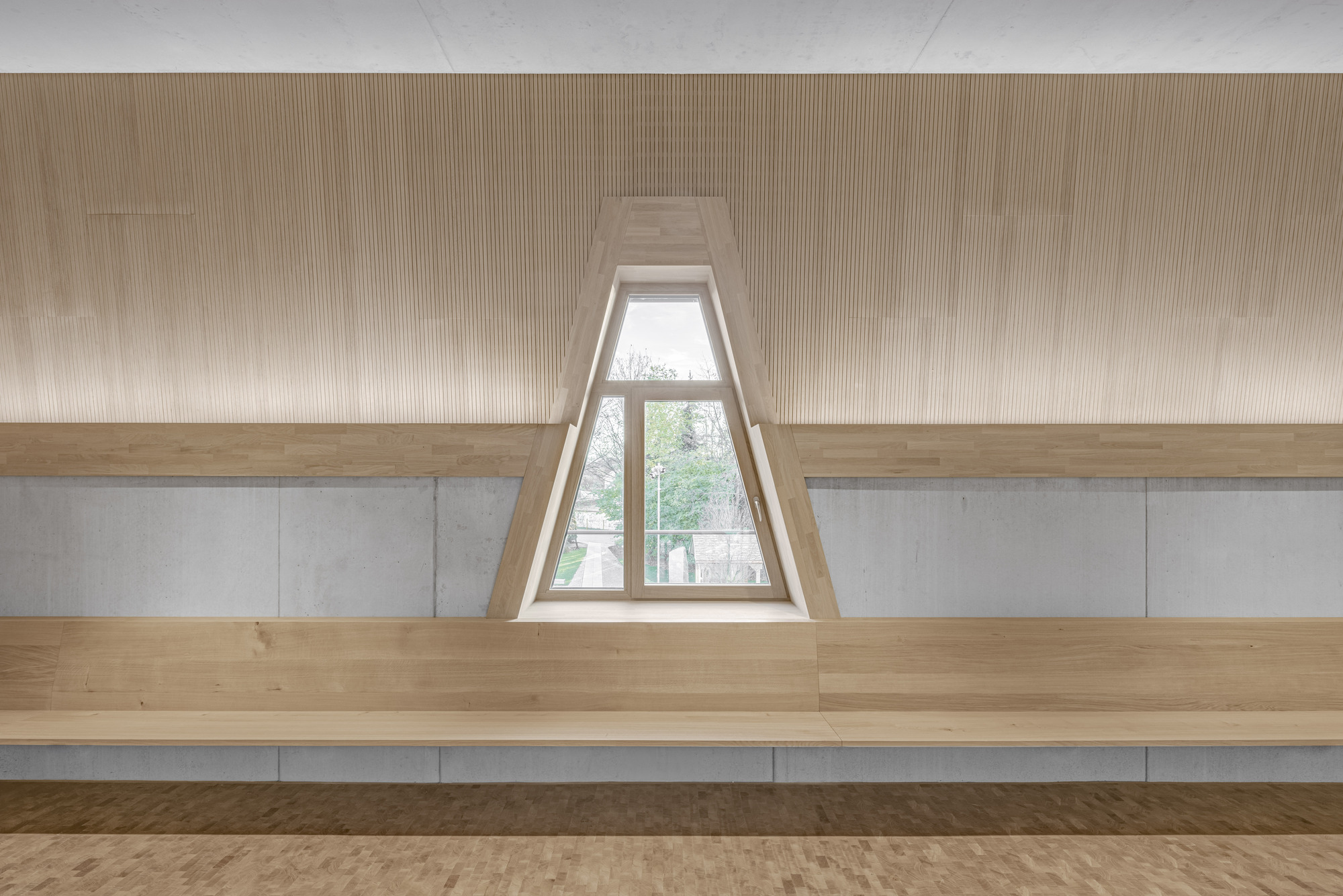 Timber bench seating along a concrete wall with a recessed triangular window framing trees outside