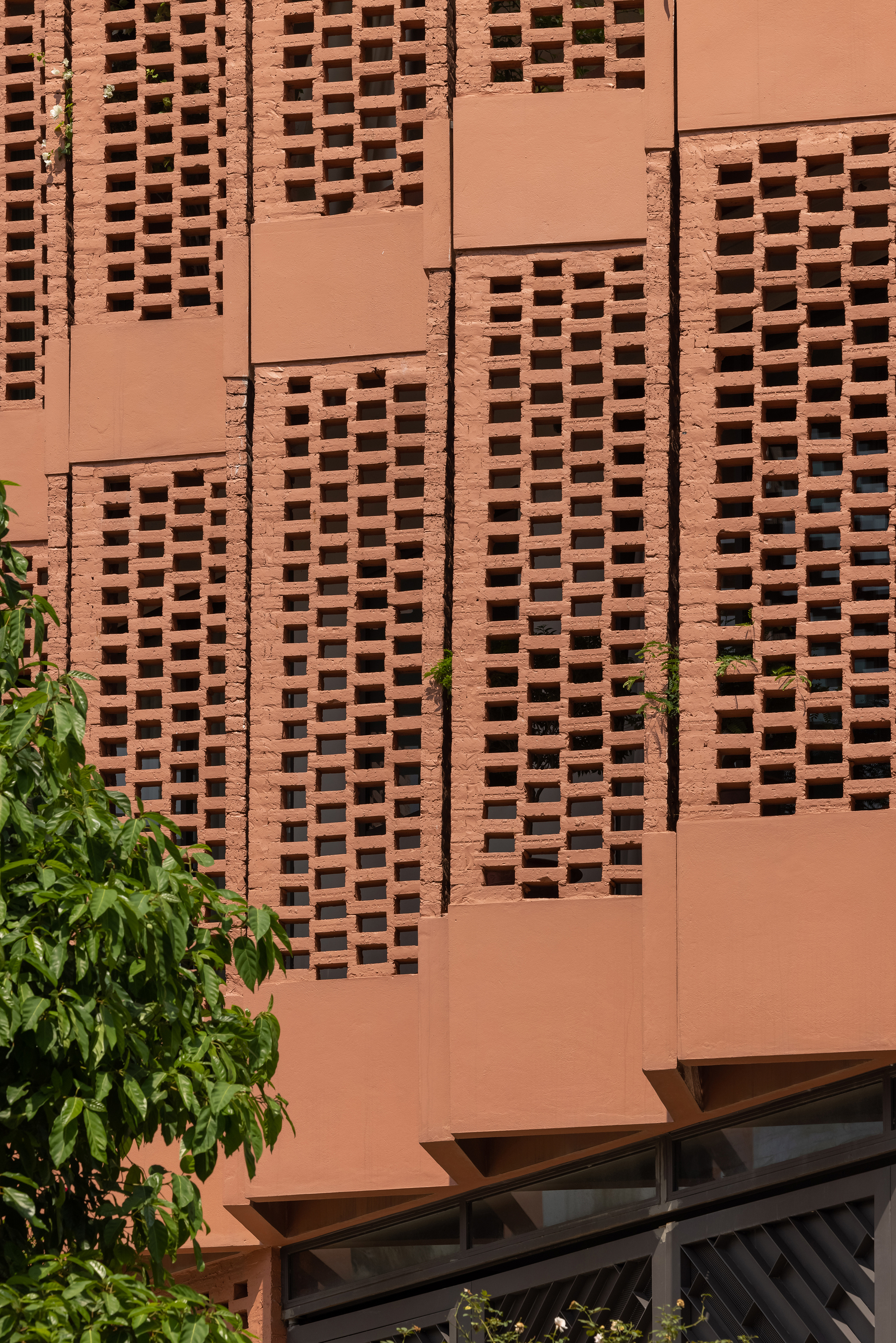Close-up of the terracotta screen wall with rectangular perforations and exposed cladding joints beside green leaves