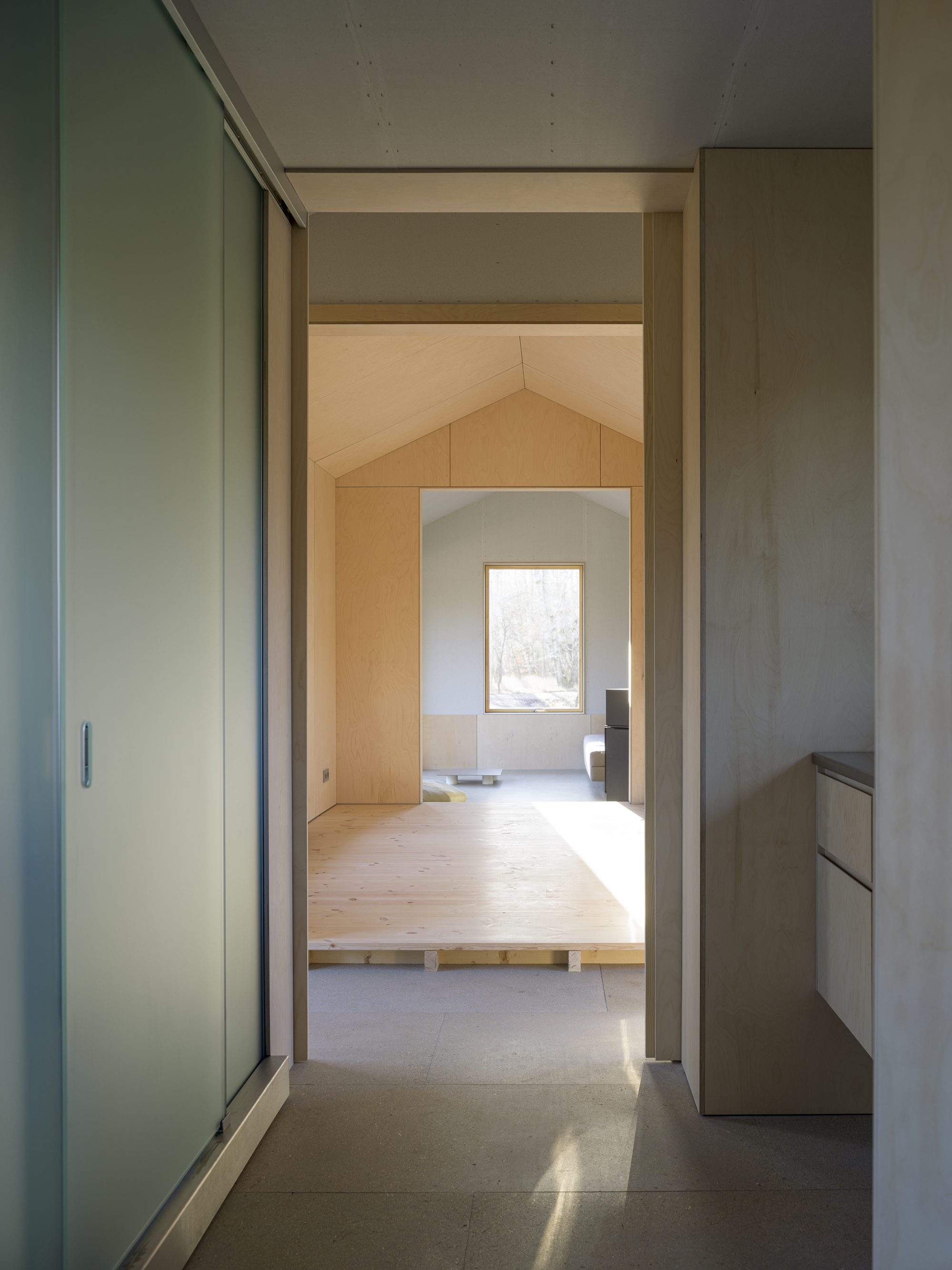 View through doorway showing sequence of plywood-lined rooms with pitched ceilings and natural light