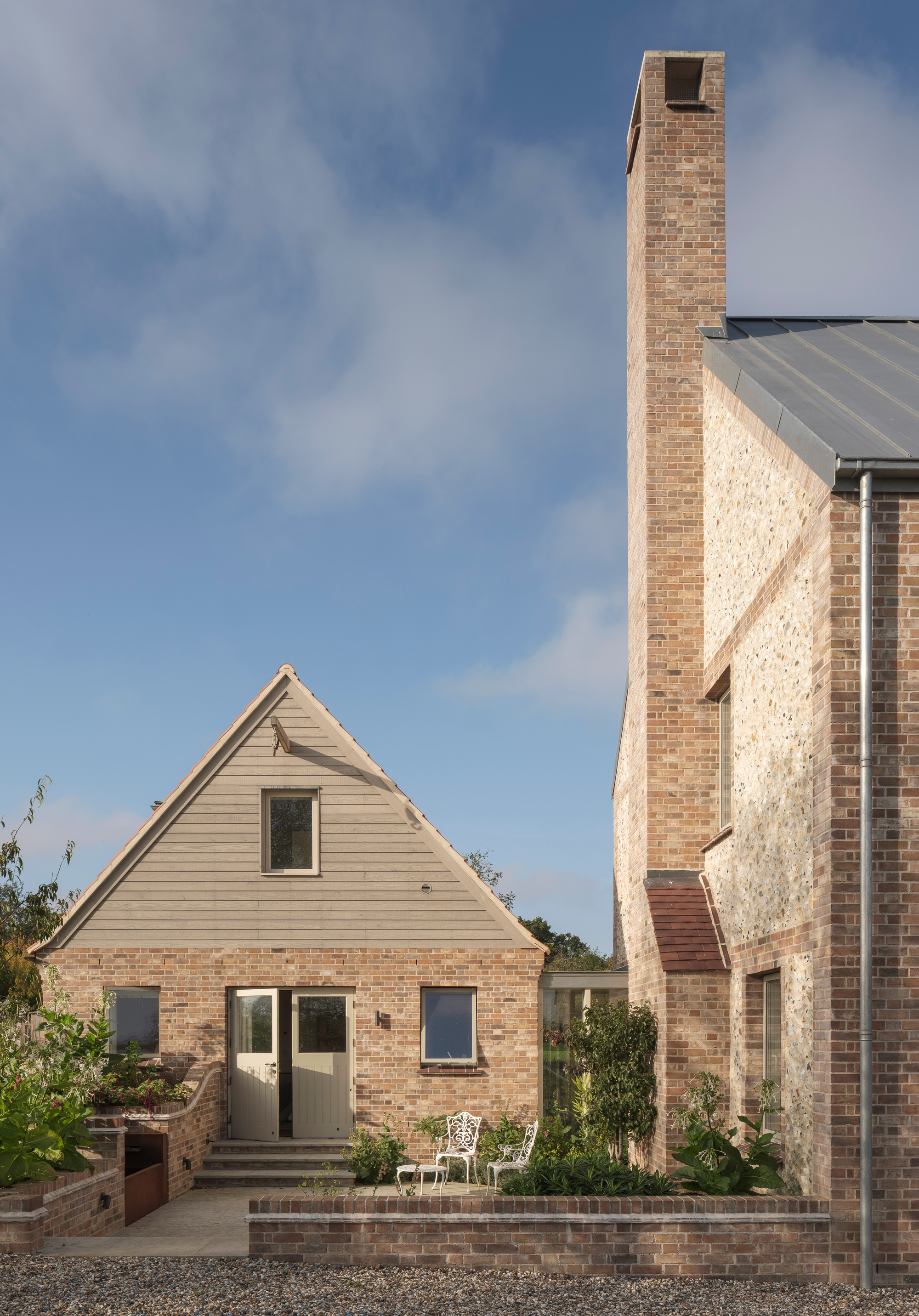Brick-paved terrace between a pale timber-clad gable and tall stone chimney with planted beds