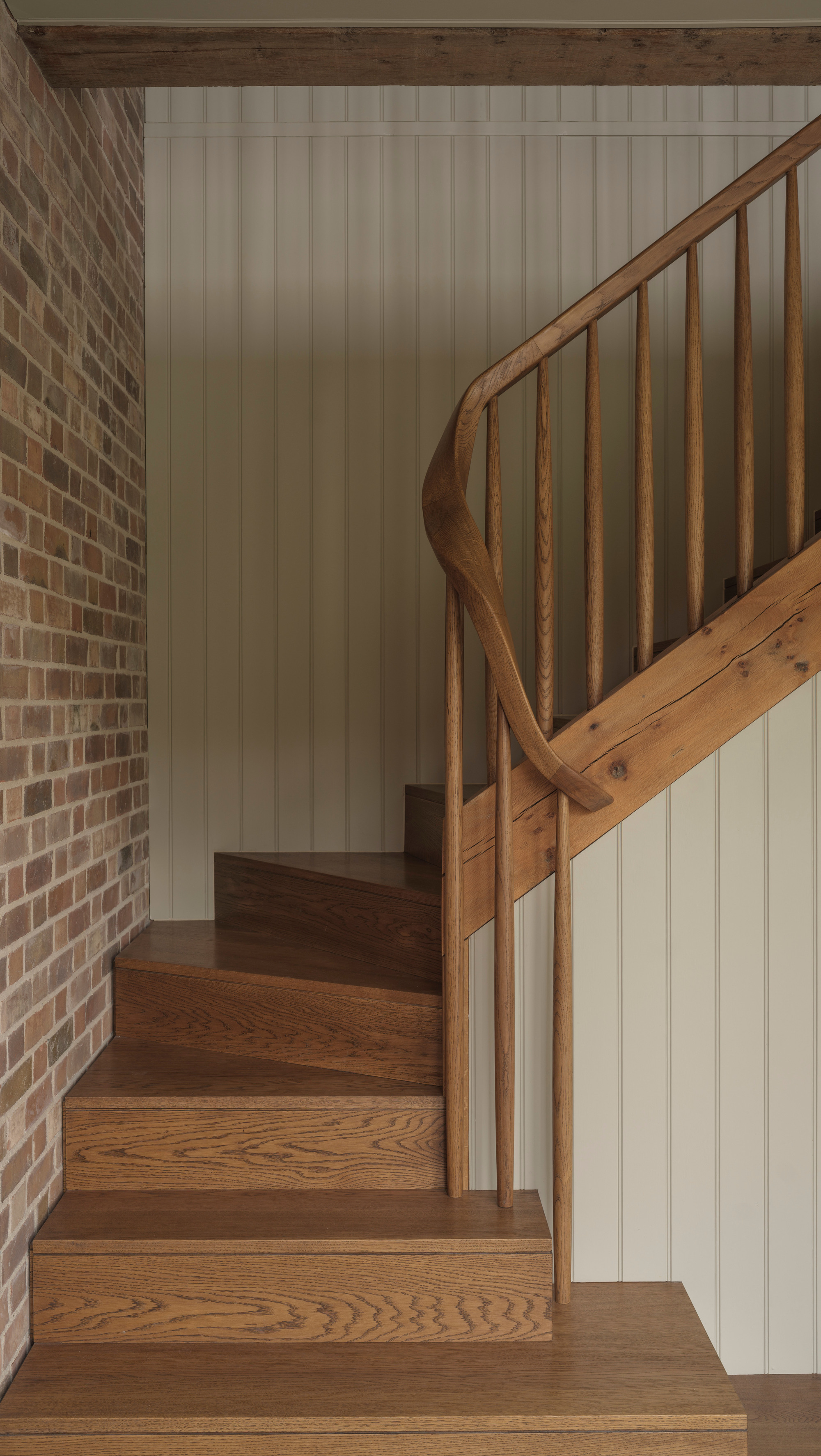 Curved timber staircase rising between brick wall and painted vertical paneling under exposed beam