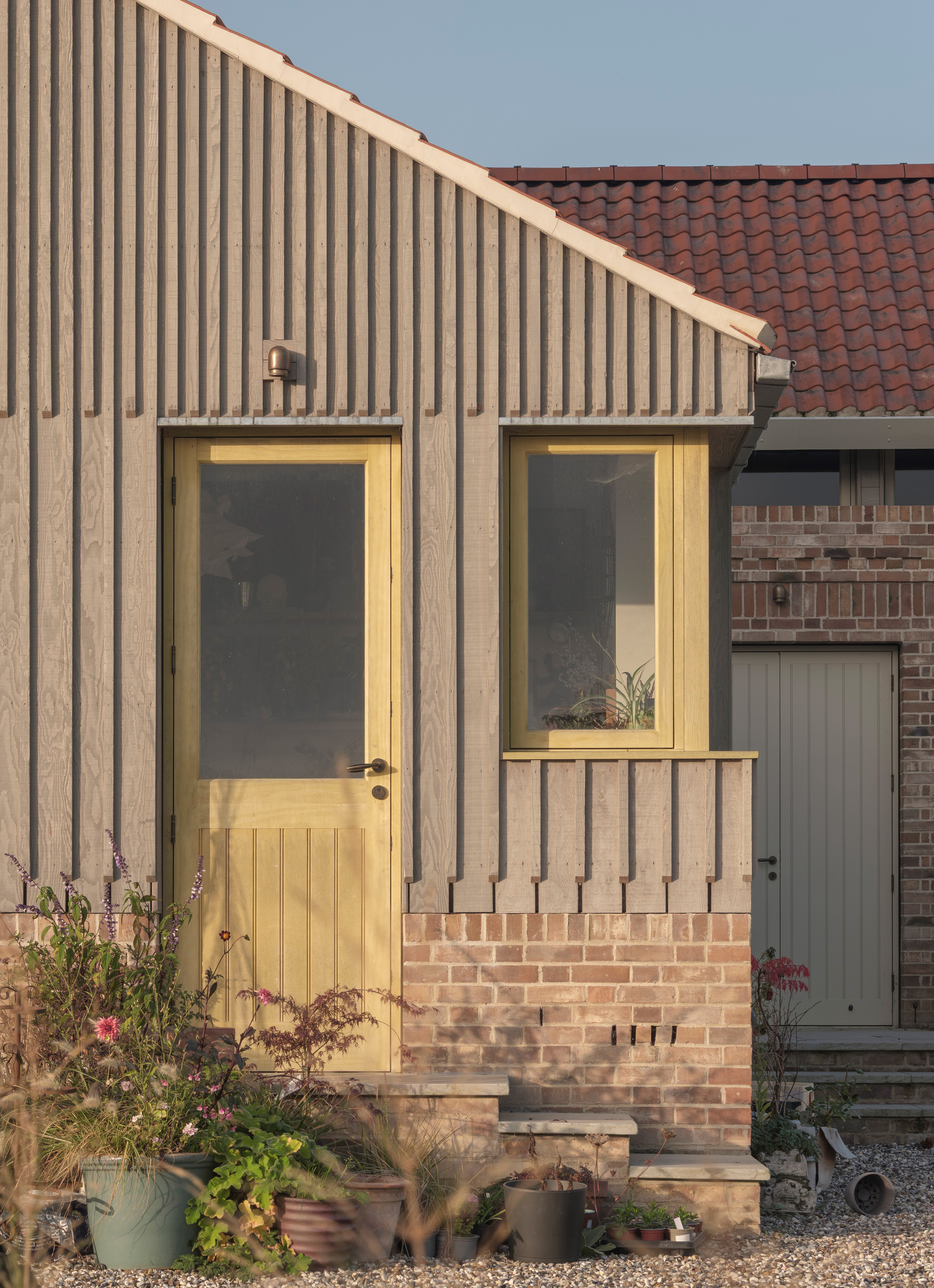 Entrance facade with timber door and window set in vertical wood cladding above brick plinth with planted bed