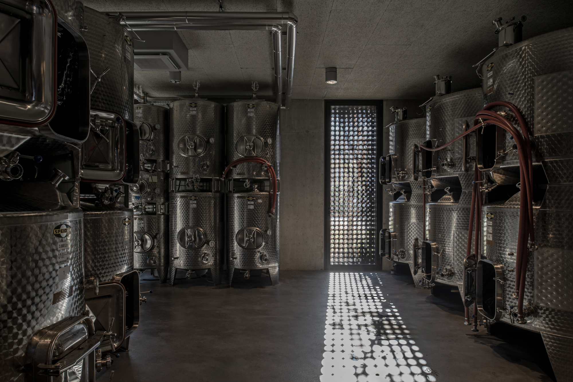 Stainless steel fermentation tanks flanking a corridor with dappled light from a perforated wall