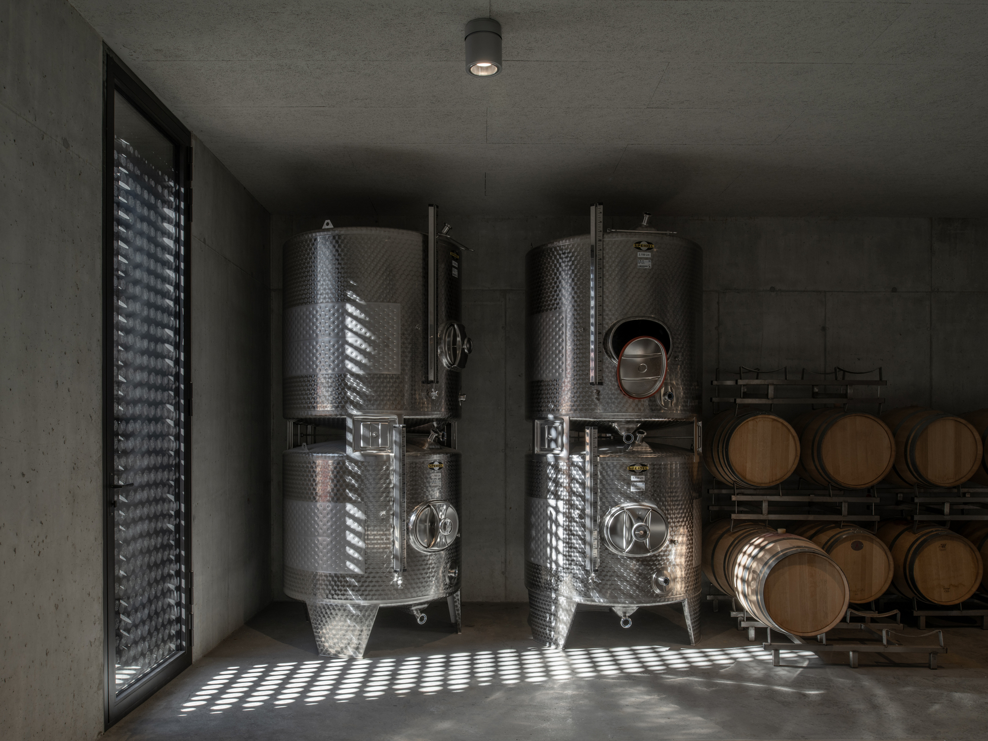 Stainless steel fermentation tanks and oak barrels in a concrete winery room with dappled light