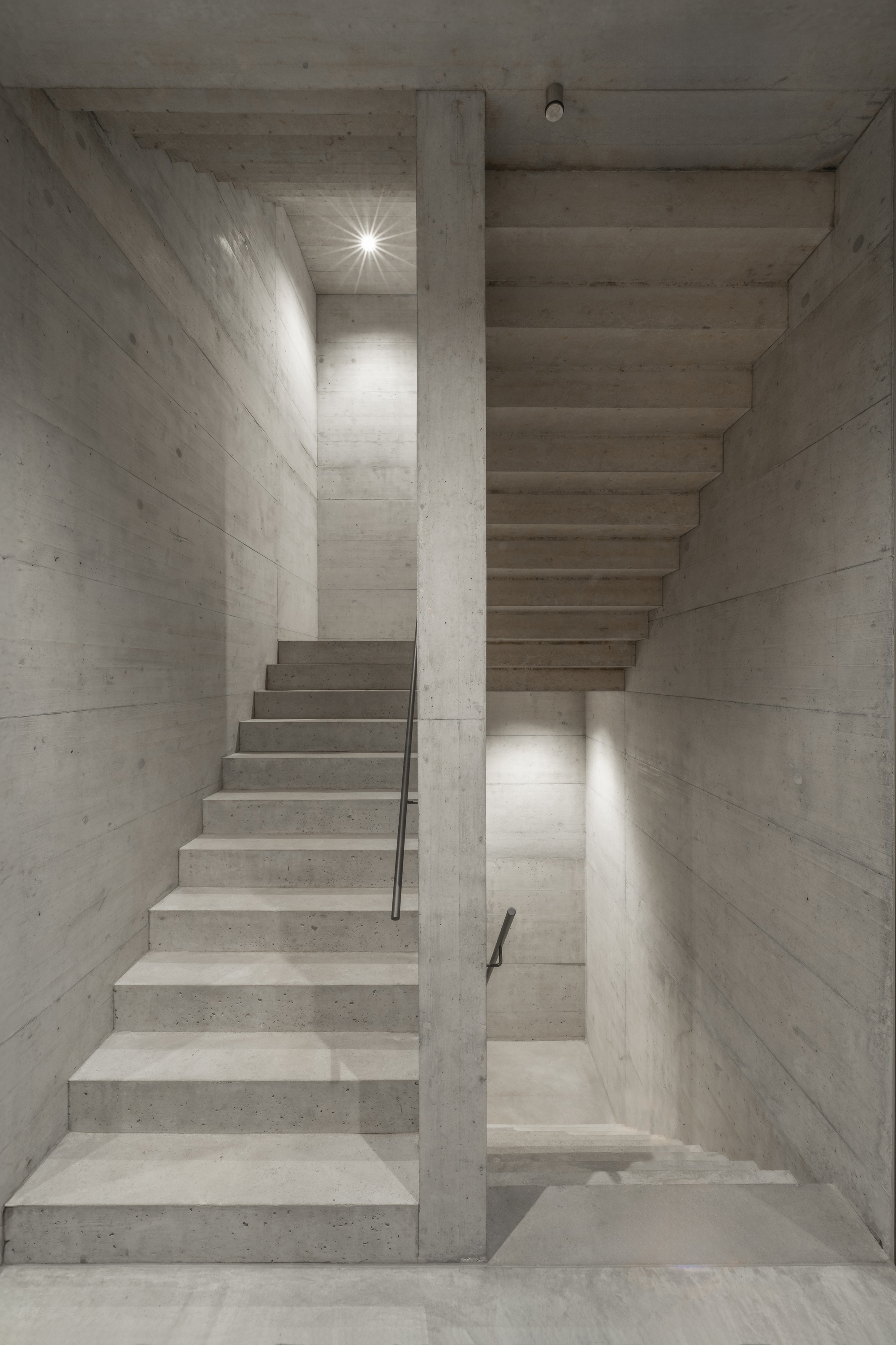 Concrete staircase wrapping around a central column with wall sconces illuminating the board-formed surfaces