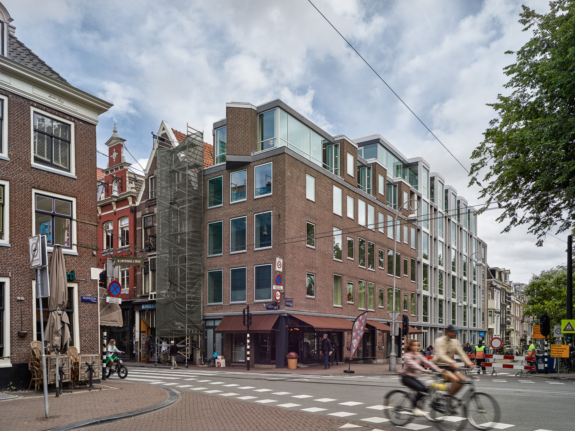Street view of the mixed brick and glass facade with cyclists passing under cloudy skies