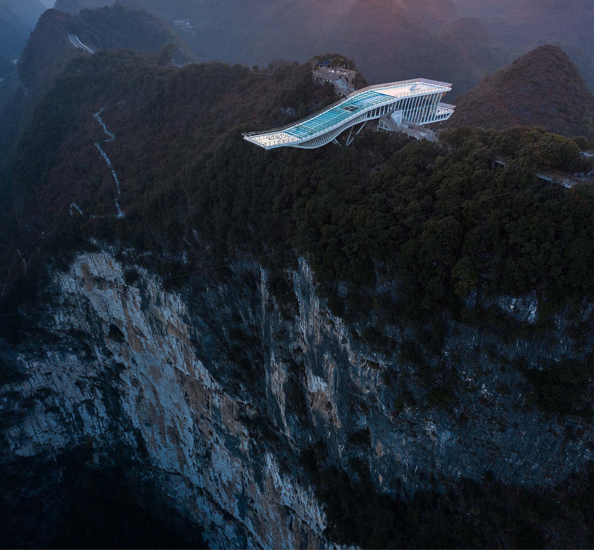 Aerial view of the wing-shaped glass platform perched on a clifftop at dusk with mist